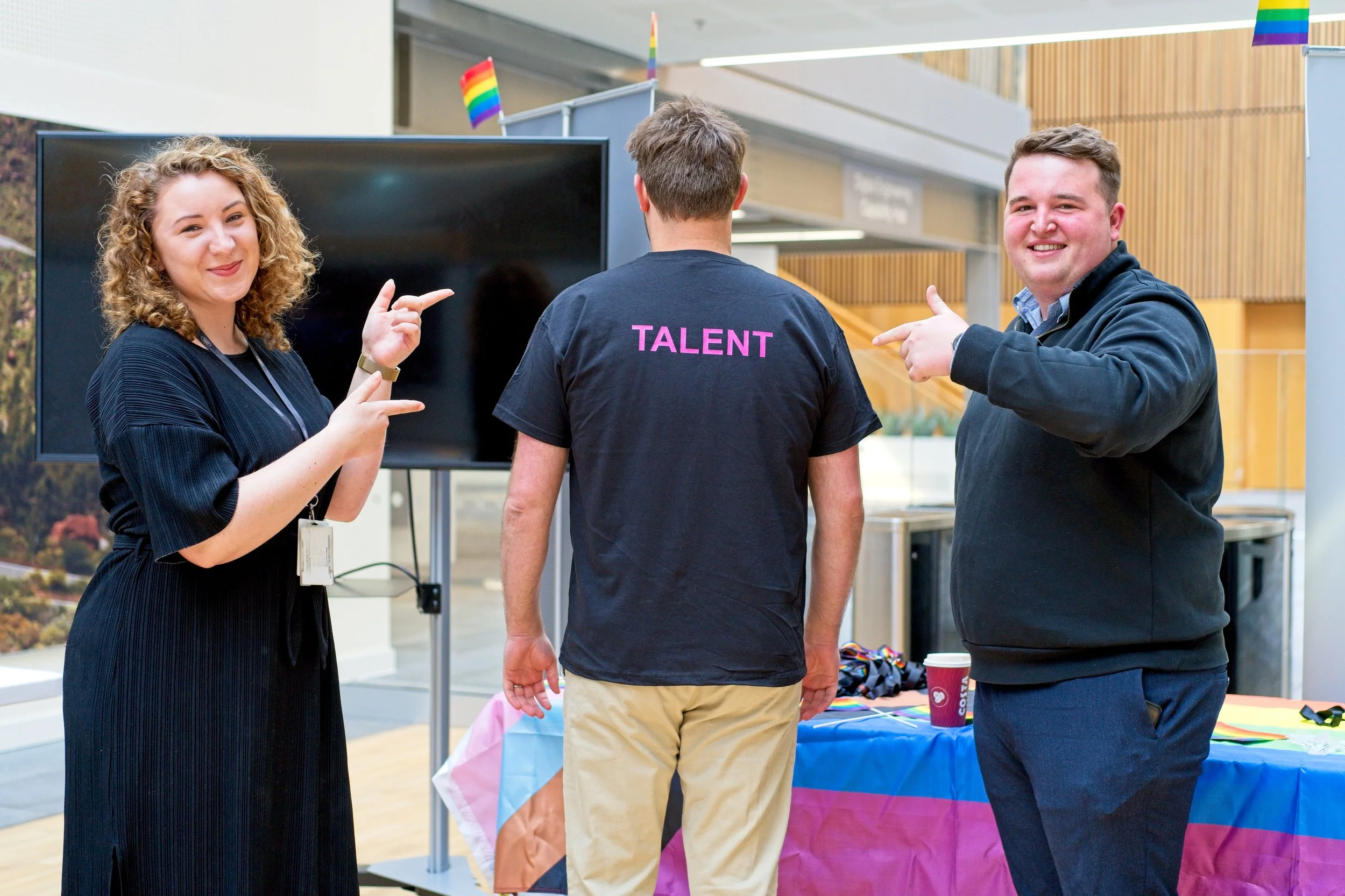 Three people at a pride event, two men and a woman, standing around a table with rainbow decor. The man in the center has his back to the camera and wears a black T-shirt with the word 'TALENT' written in pink on the back. The woman on the left and t