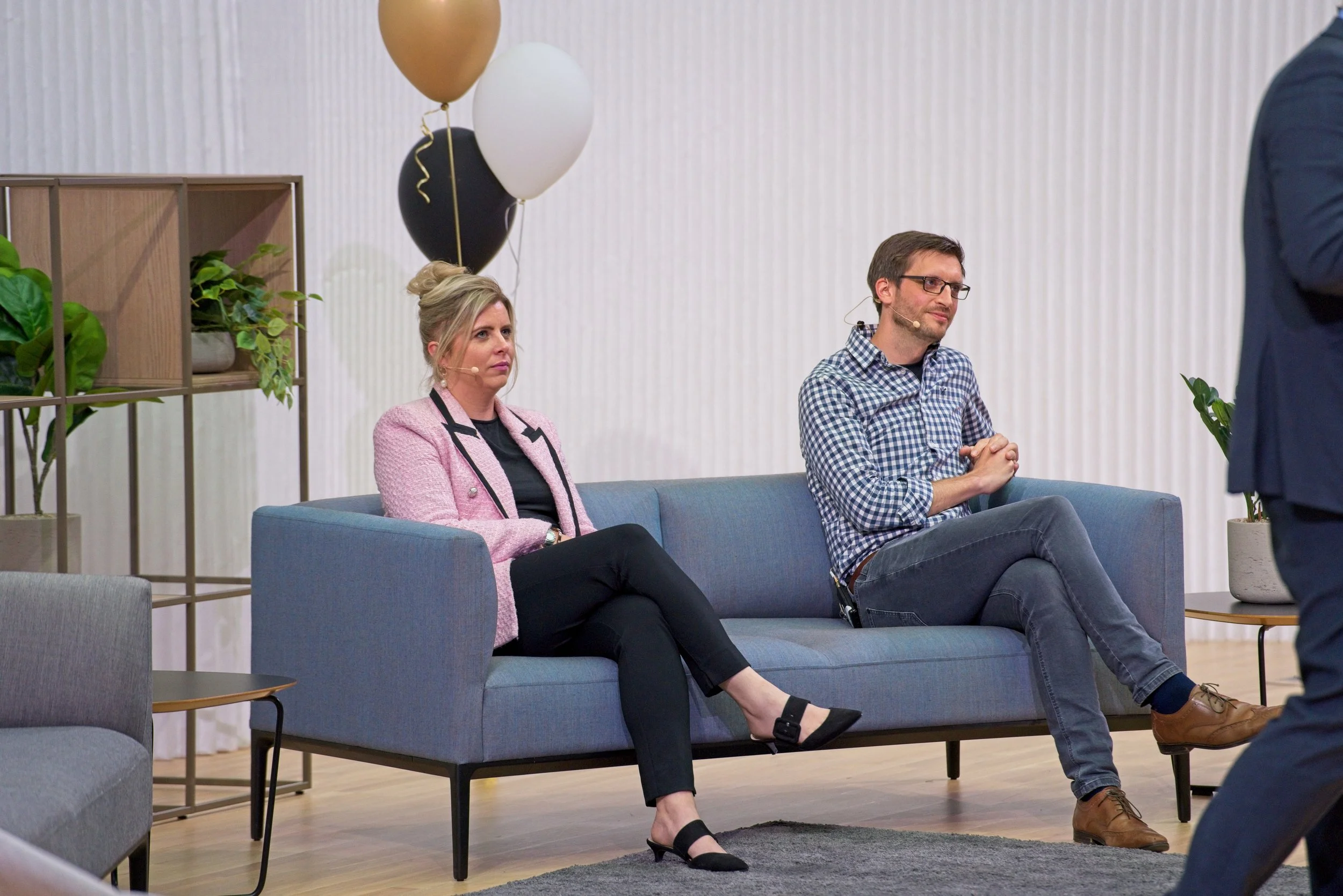 Two people sitting on a blue sofa, attending a presentation or seminar, with white and black balloons in the background. Event Photography by photographer based in Southam, Warwickshire.