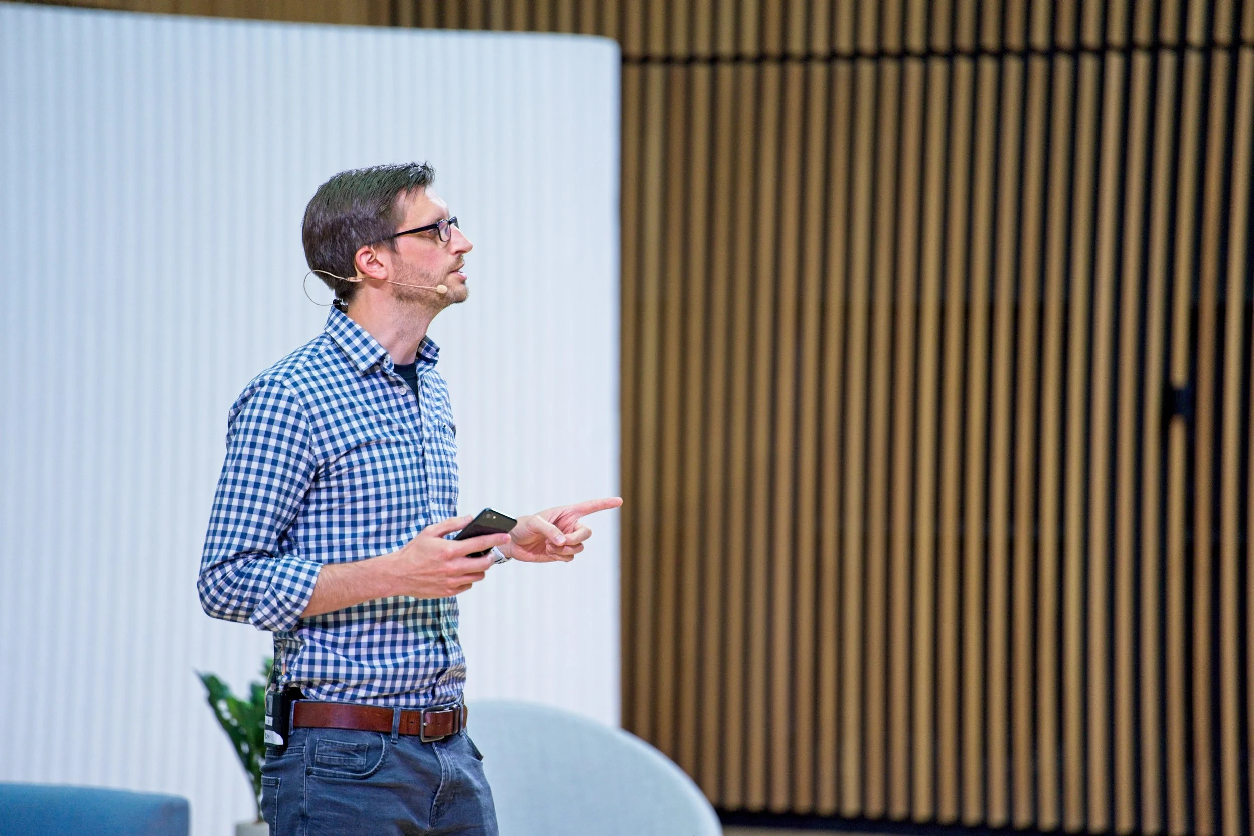 Man in checkered shirt and glasses giving a presentation, holding a remote control, in a modern conference room. Event Photography by photographer based in Southam, Warwickshire.