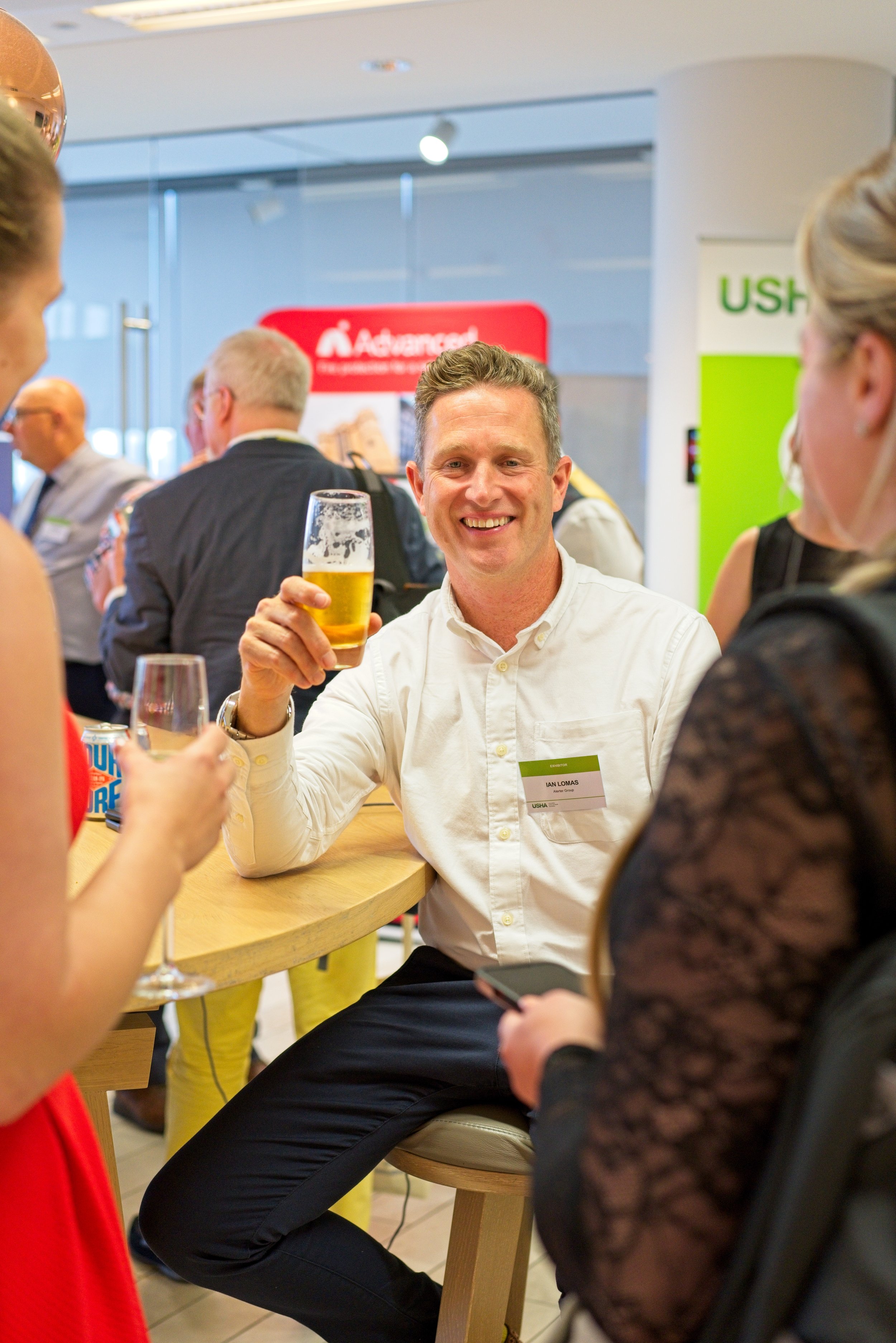A man smiling and holding a glass of beer while sitting at a round table with two women, at a social event or conference. Event Photography by photographer based in Southam, Warwickshire.