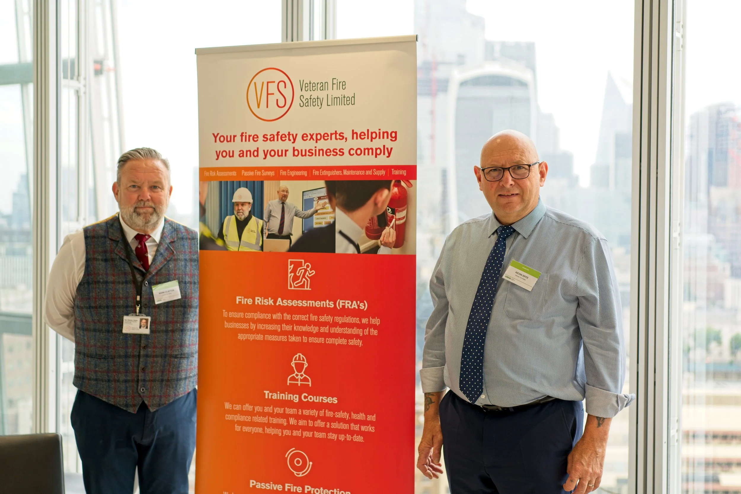Two men standing next to a red and white banner with fire safety information at a professional event in a high-rise building with large windows, cityscape view in the background. Event Photography by photographer based in Southam, Warwickshire.