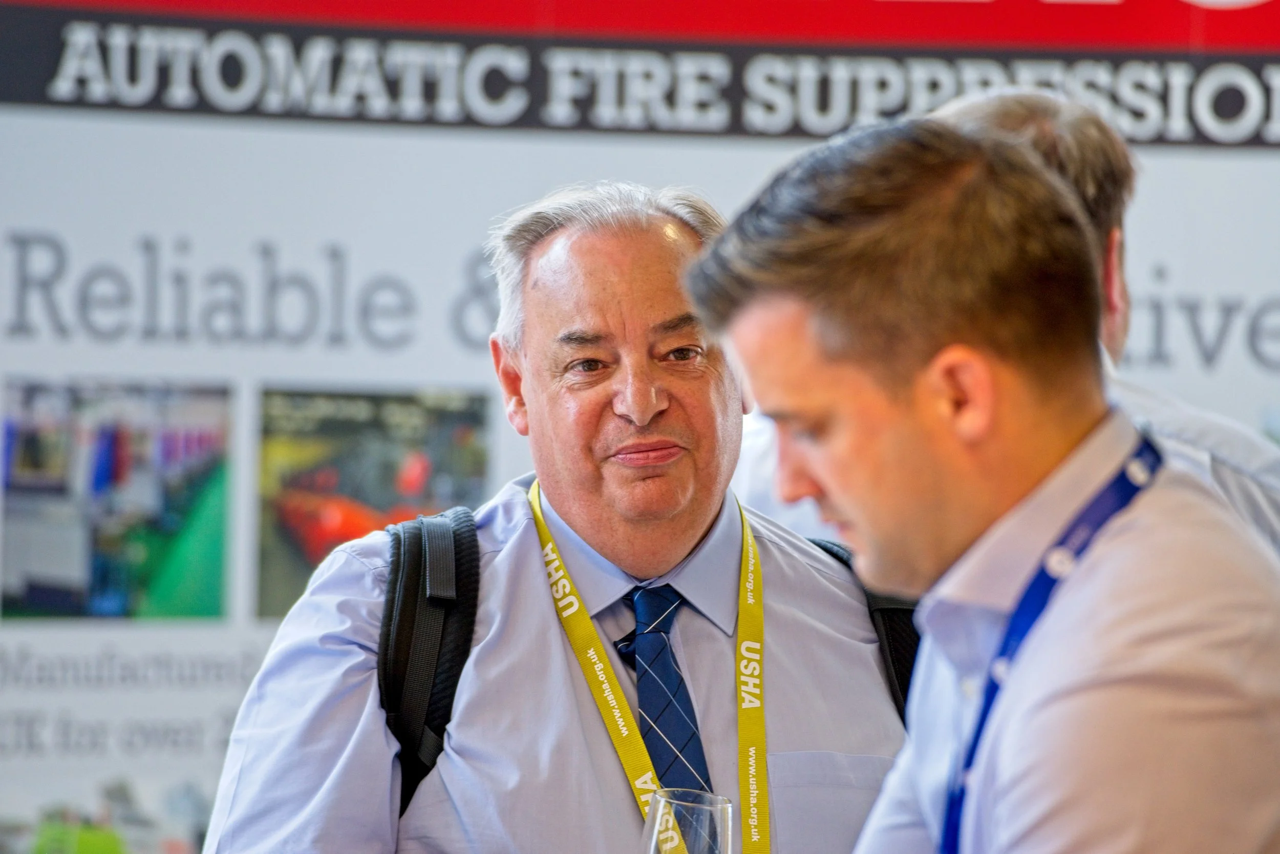 Two men in business attire attending a conference, with the focus on an older man with gray hair and a blue tie. The background features a banner with the words "AUTOMATIC FIRE SUPPRESSION" and some images. Event Photography Warwickshire