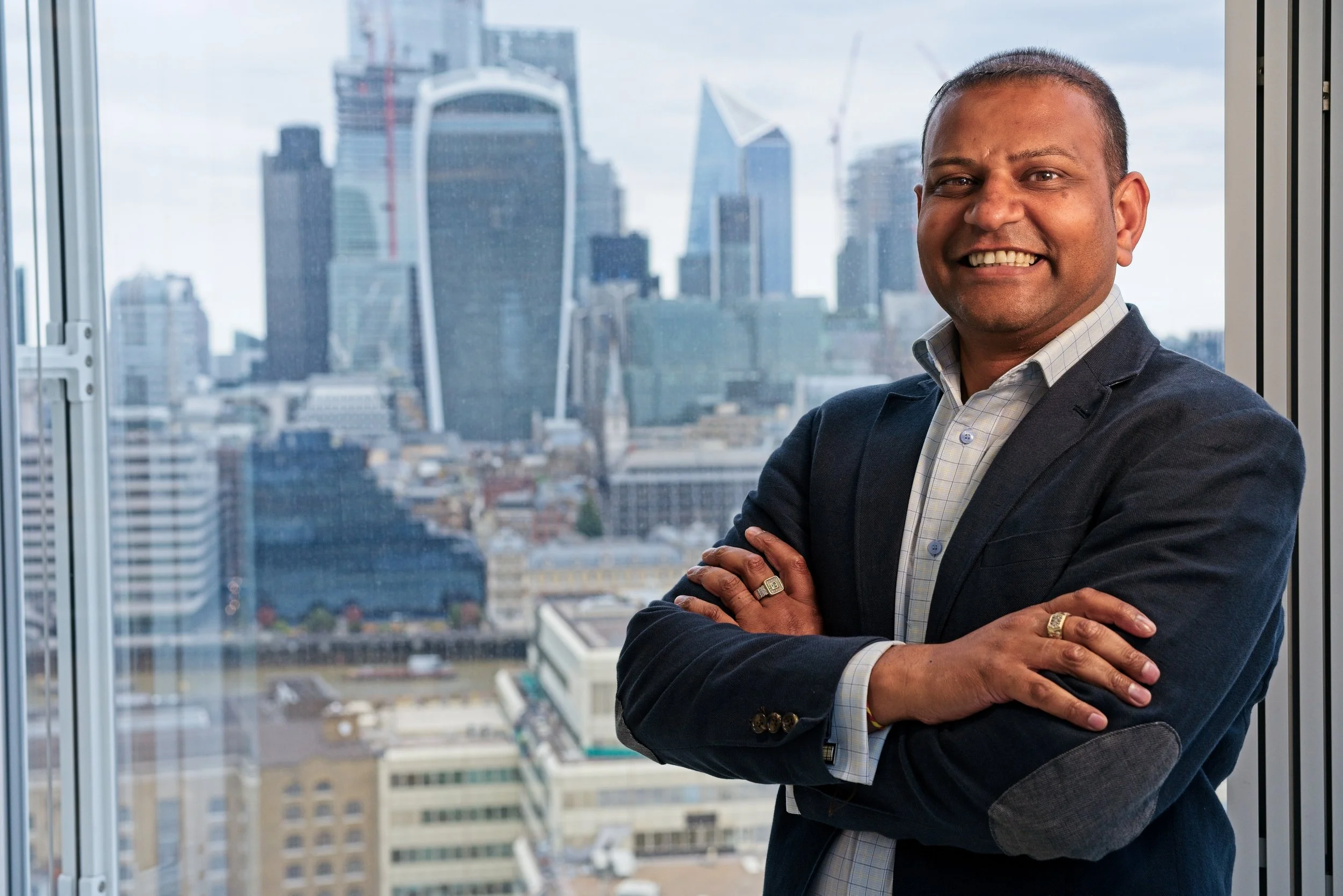 A smiling man in a business suit standing with arms crossed in front of a window overlooking a city skyline. Event Photography by photographer based in Southam, Warwickshire.