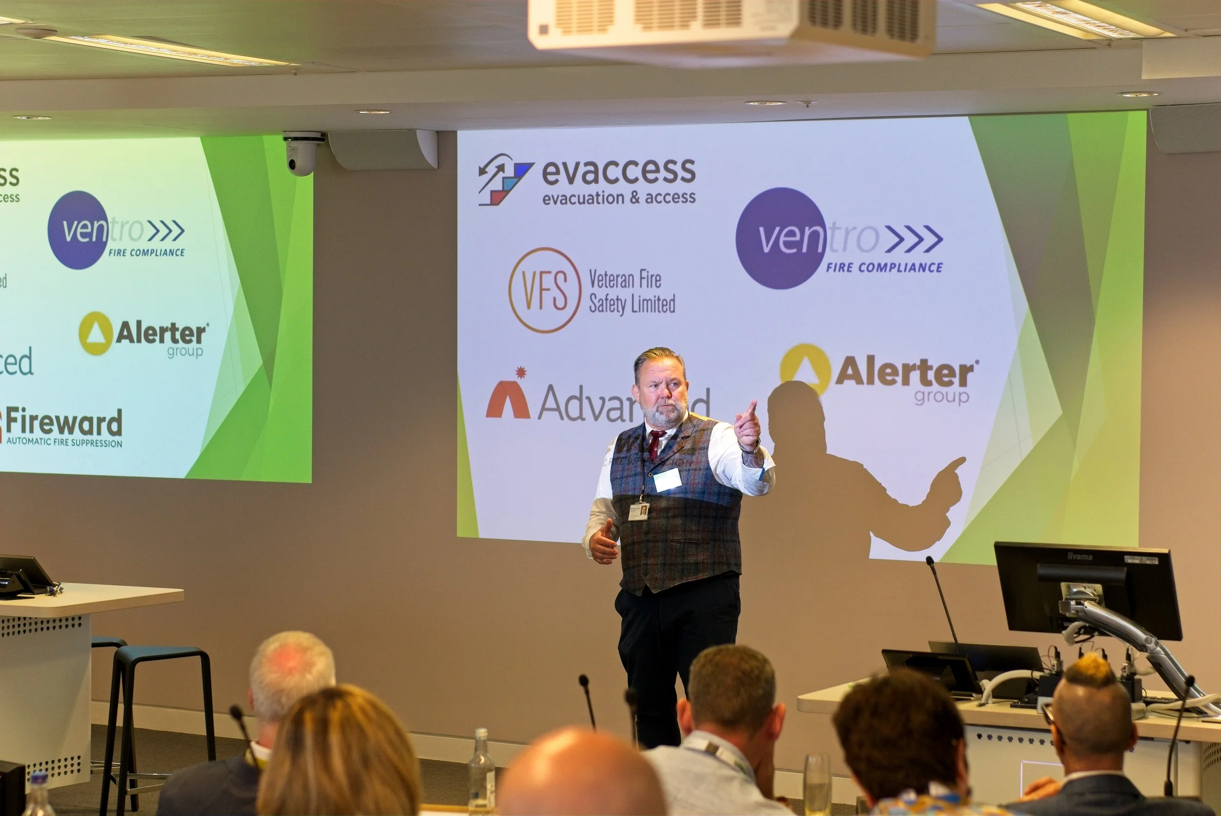 A man giving a presentation at a conference with a slide projected behind him displaying logos of various fire safety and evacuation companies, including Evaccess, Ventro, Veteran Fire Safety Limited, Alerter group, Advanced, and Fireward, in a profe