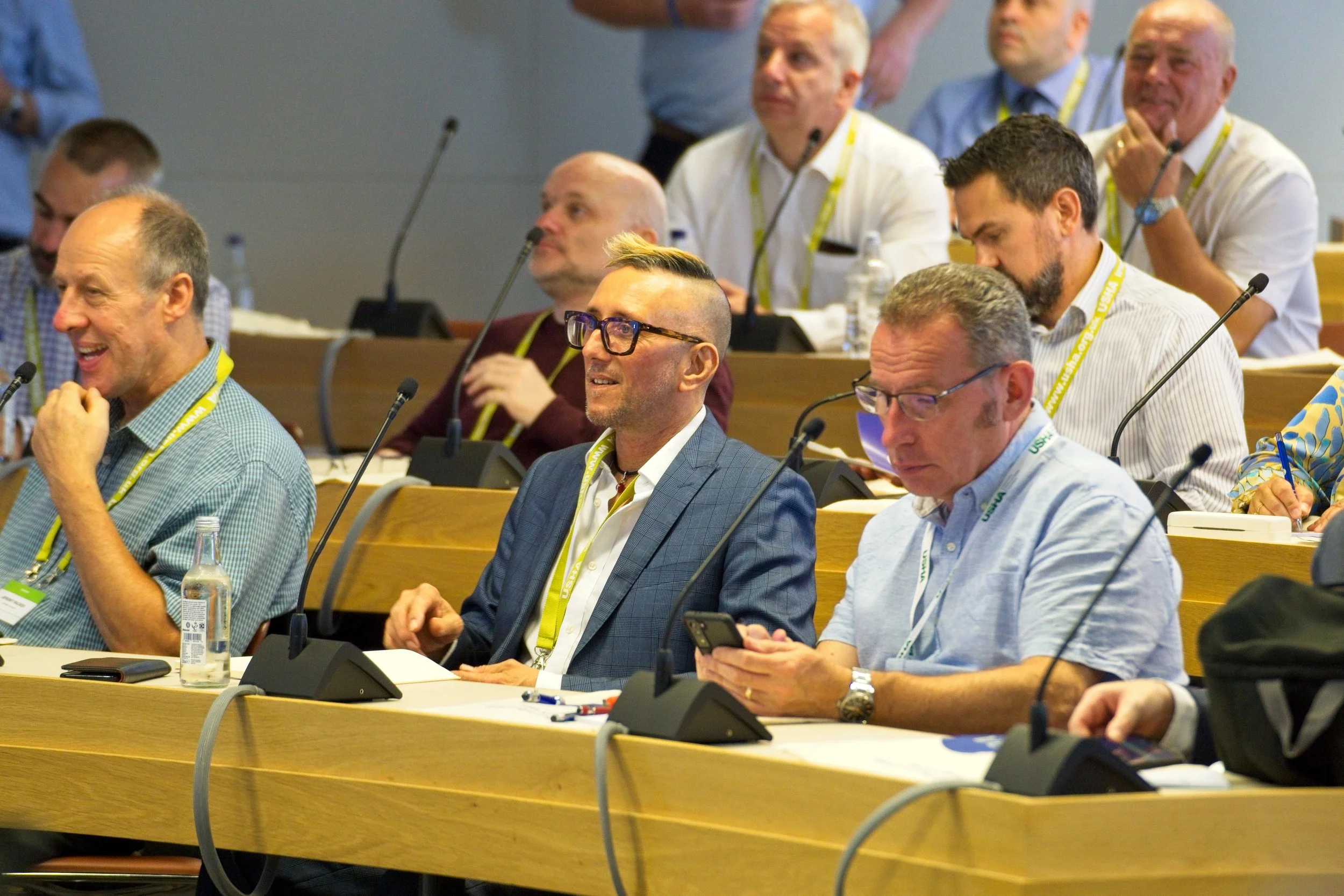 Attendees at a conference room listening to a presentation, some are looking at their phones, others are smiling or taking notes. Event Photography by photographer based in Southam, Warwickshire.