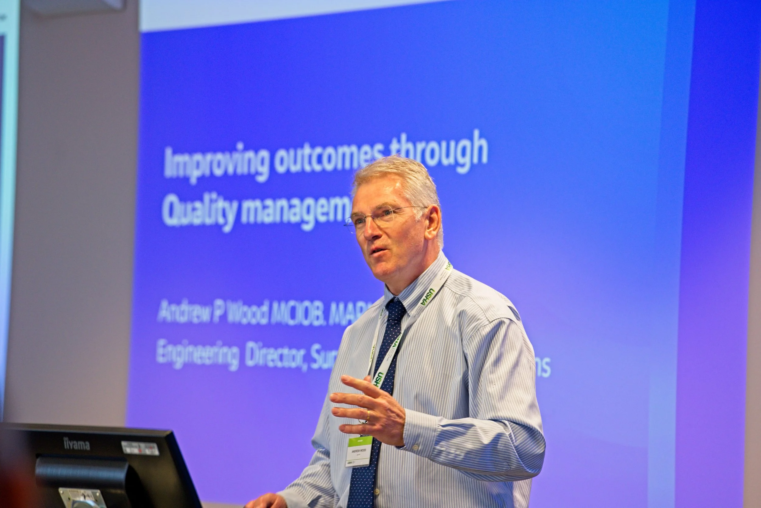A man with glasses and gray hair in a striped shirt and tie giving a presentation in front of a blue slide that reads 'Improving outcomes through Quality management'. Event Photography by photographer based in Southam, Warwickshire.
