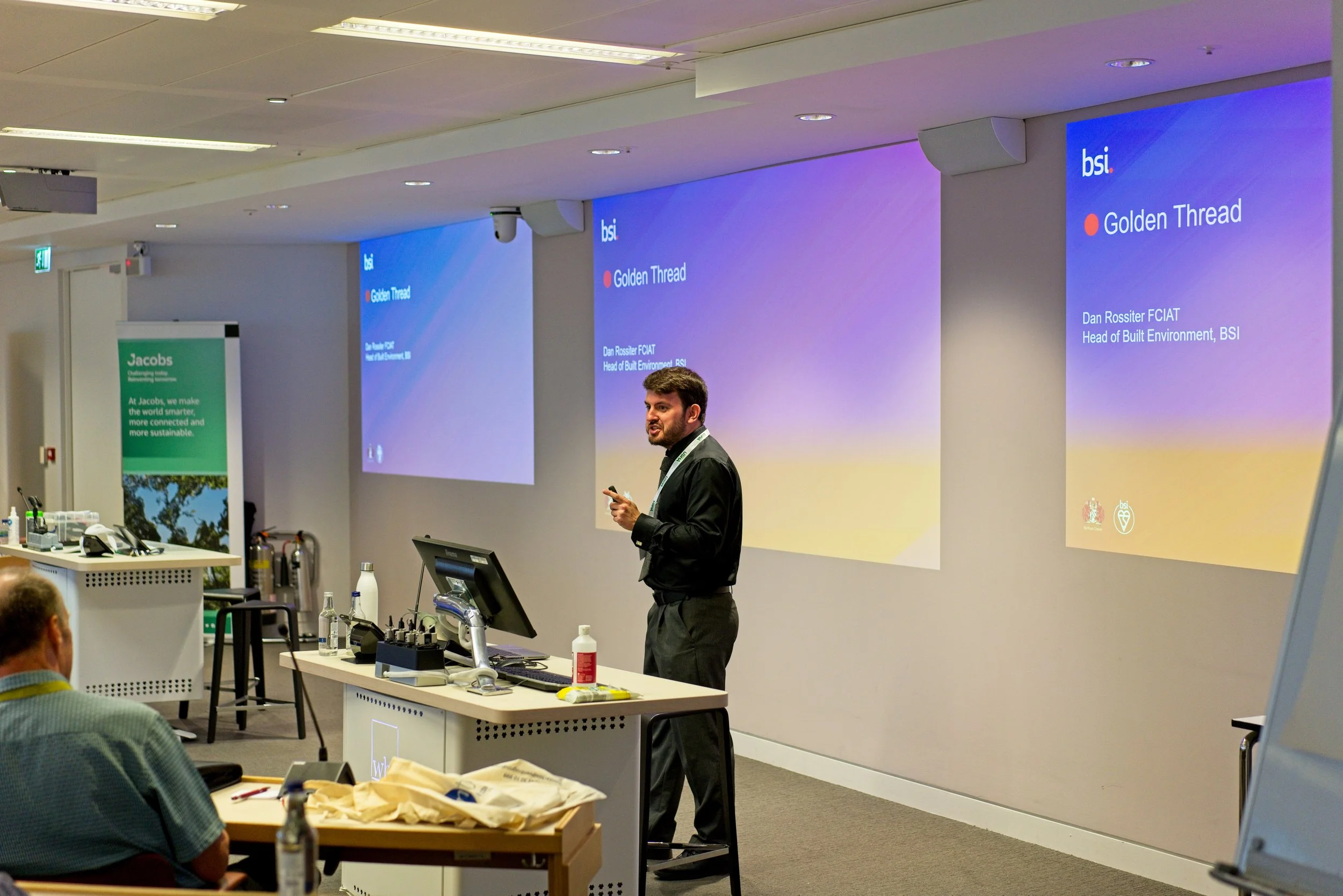 A man giving a presentation in a conference room with multiple screens displaying a slide titled 'Golden Thread.' Event Photography by photographer based in Southam, Warwickshire.