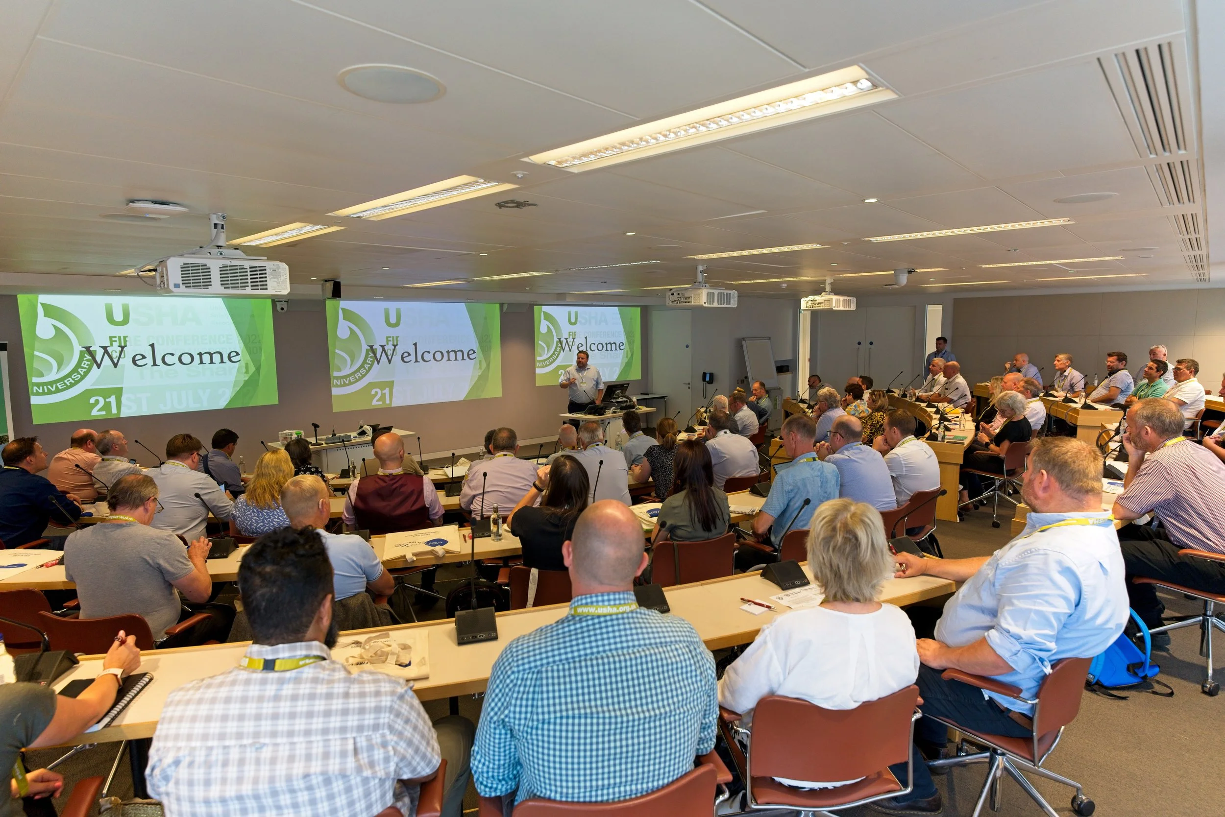 A large conference room filled with attendees sitting at long tables, listening to a speaker at the front. There are three screens displaying the welcome message for the USHA conference, dated July 21. Event Photography Warwickshire.