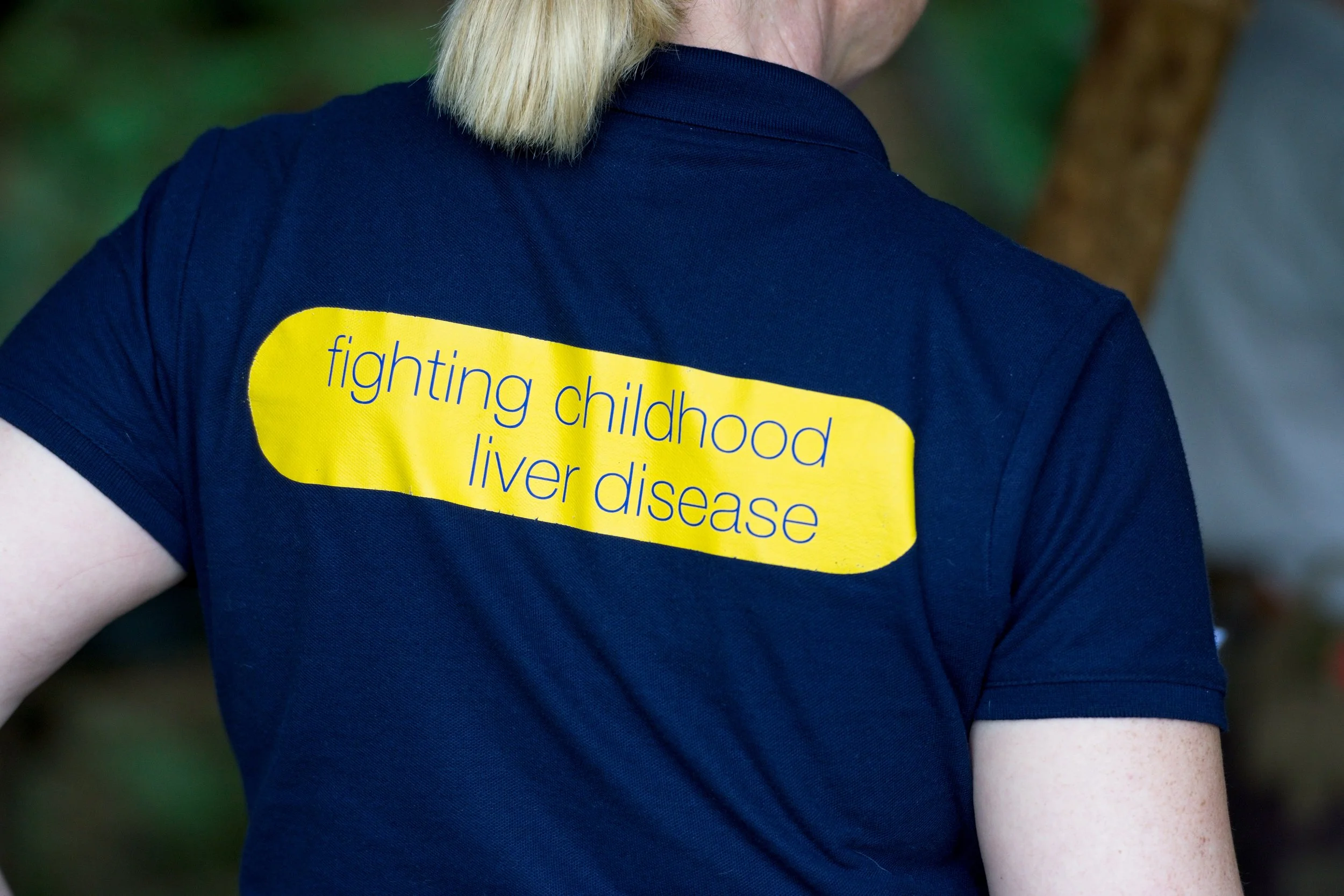 Back of person wearing dark blue T-shirt with yellow patch stating 'fighting childhood liver disease' in purple text. Event Photography by photographer based in Southam, Warwickshire.