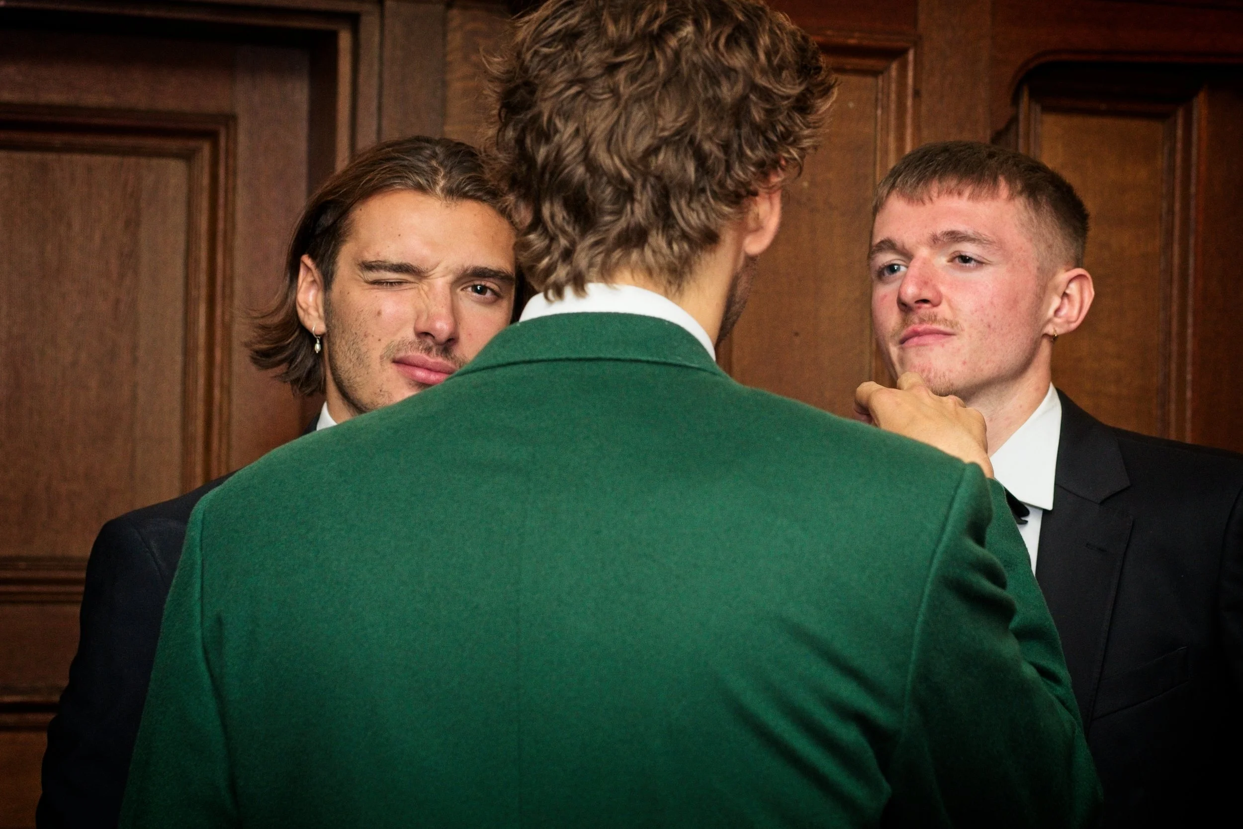 Two men in tuxedos and one man in a green jacket engage in a conversation in a wood-paneled room, with one of the tuxedoed men making a playful facial expression. Event Photography by photographer based in Southam, Warwickshire.
