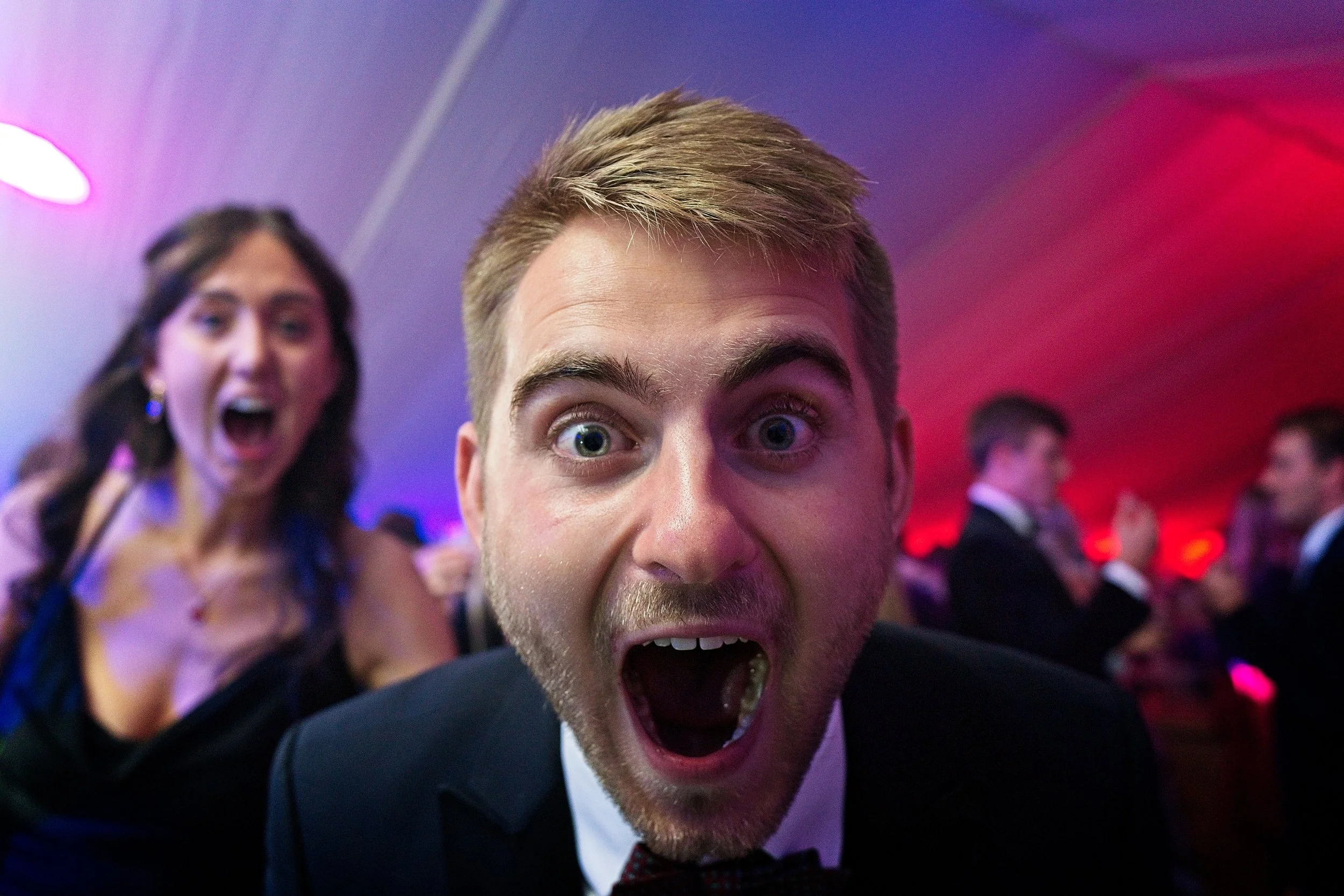 A close-up of a young man with a surprised or excited expression at a party or social event, with a woman behind him also appearing excited and others in the background. Event Photography by photographer based in Southam, Warwickshire.