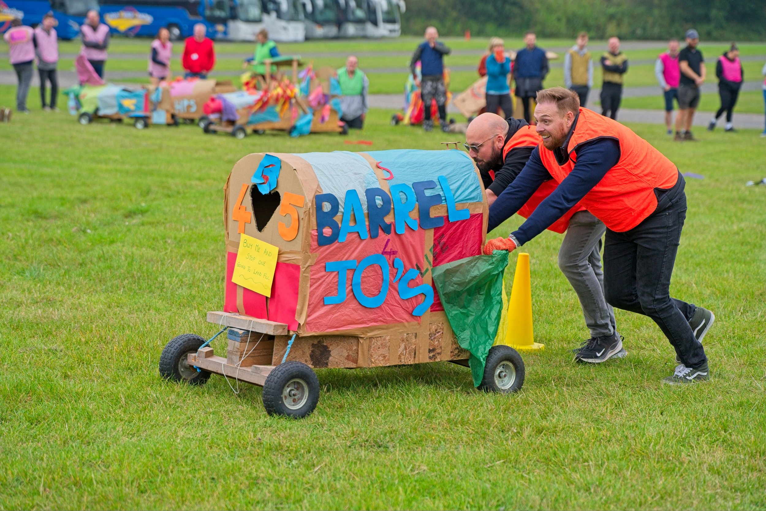 Two men in orange vests pushing a decorated cardboard container on wheels across a grassy field during an outdoor event, with others and parked buses in the background.