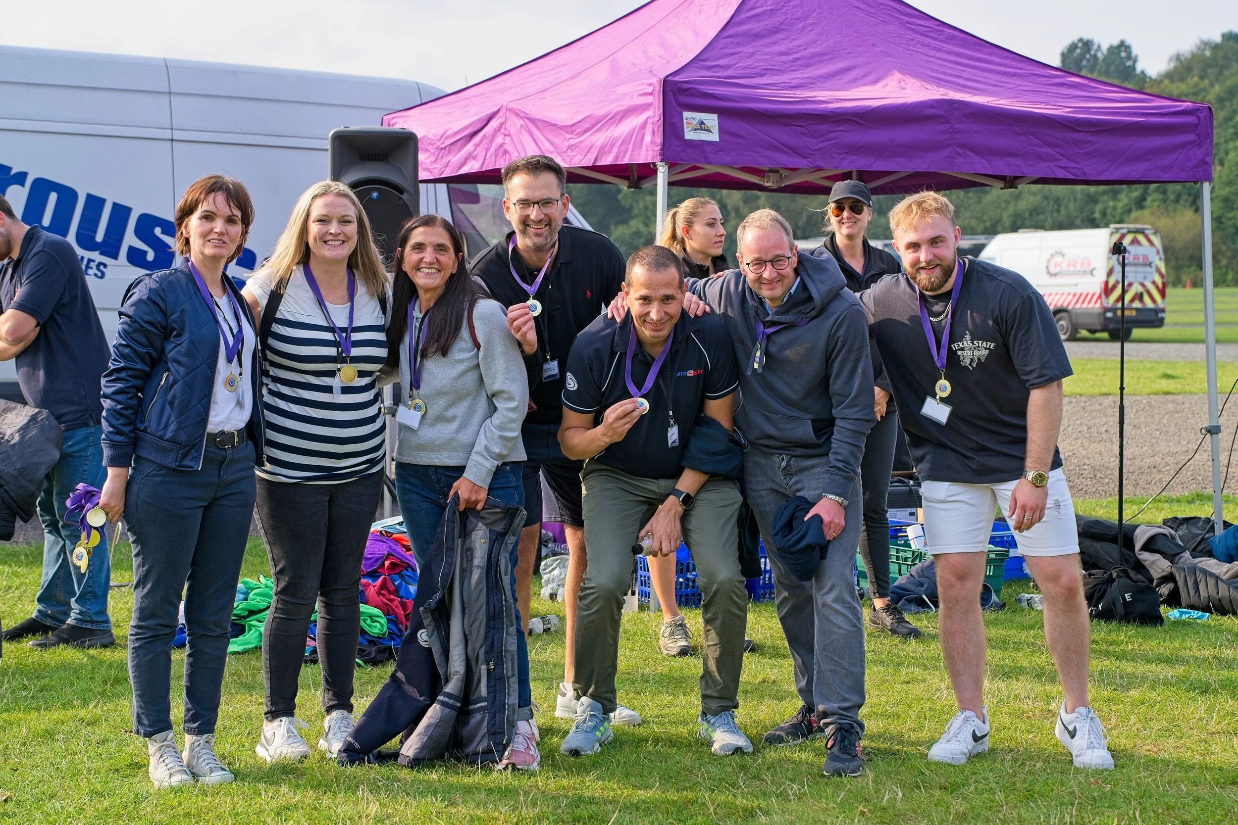 Group of nine people standing outdoors on a grassy field, smiling, with some holding medals. Behind them is a purple canopy tent and a white vehicle, with equipment laid out on the ground.