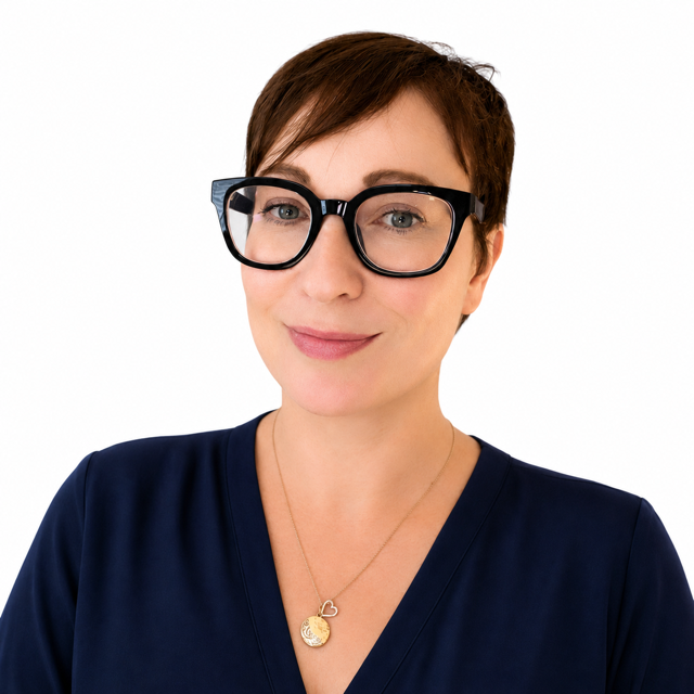A woman with short brown hair wearing large black glasses, a navy blue top, and a gold necklace with two pendants.