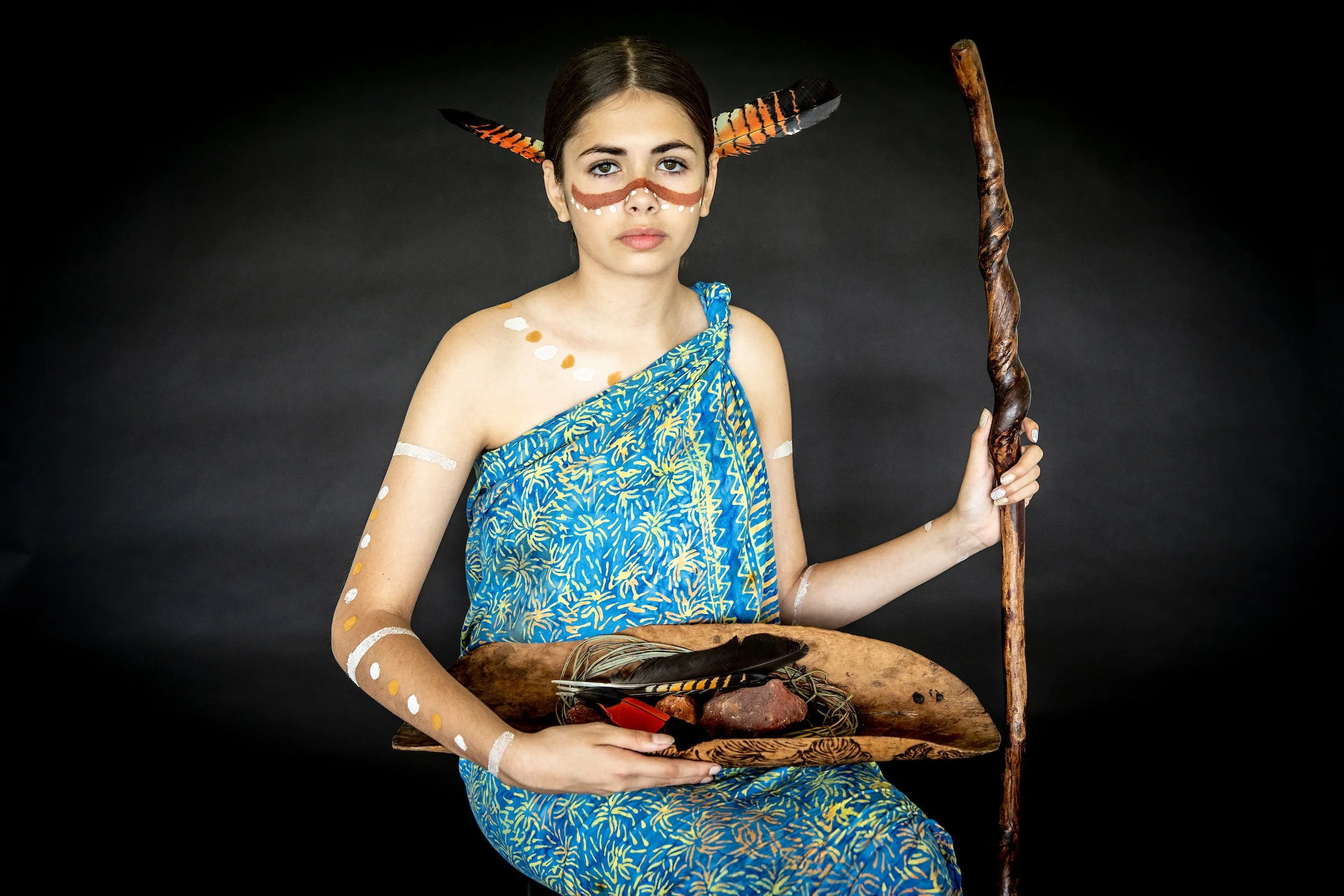 A young woman with tribal face paint, painted in brown with white dots, is dressed in blue and yellow patterned fabric, holding a wooden staff, and seated on a carved wooden chair with a black background.