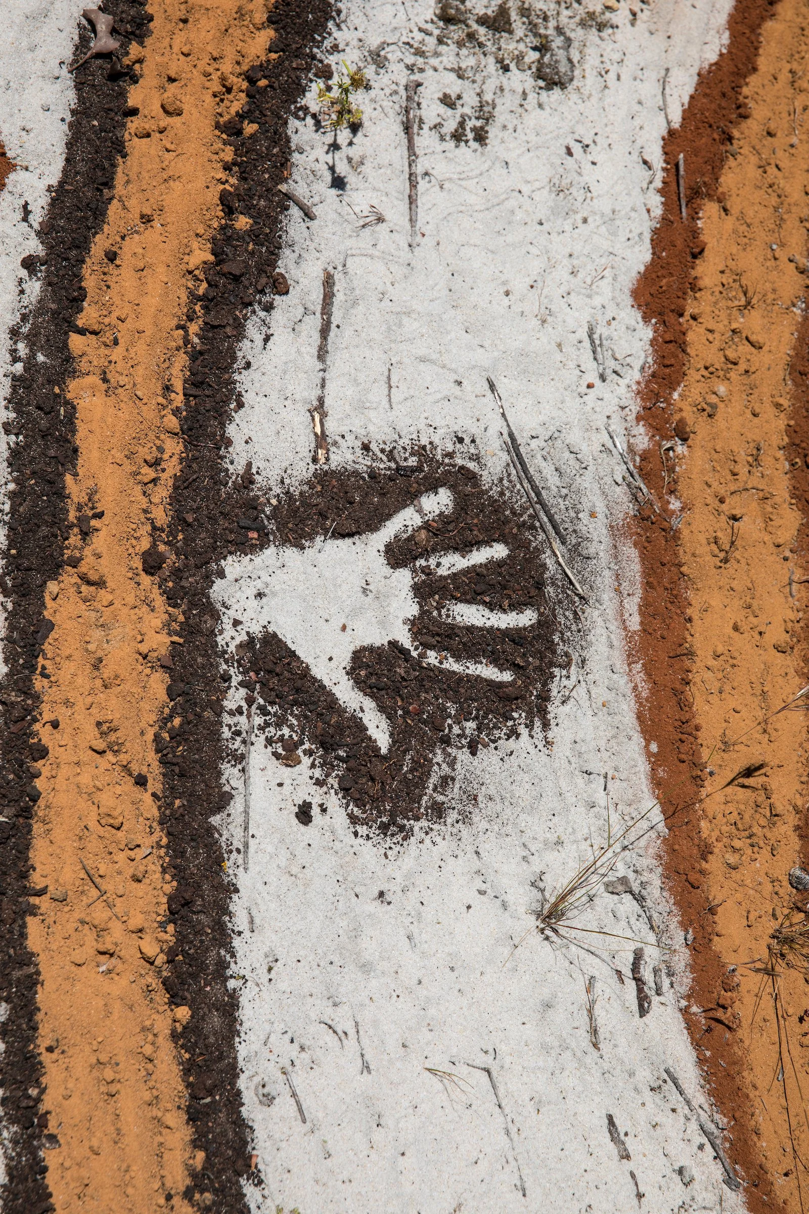 A sidewalk with a white stencil of a human handprint on the concrete, surrounded by alternating layers of orange and brown soil, with small patches of grass and twigs.