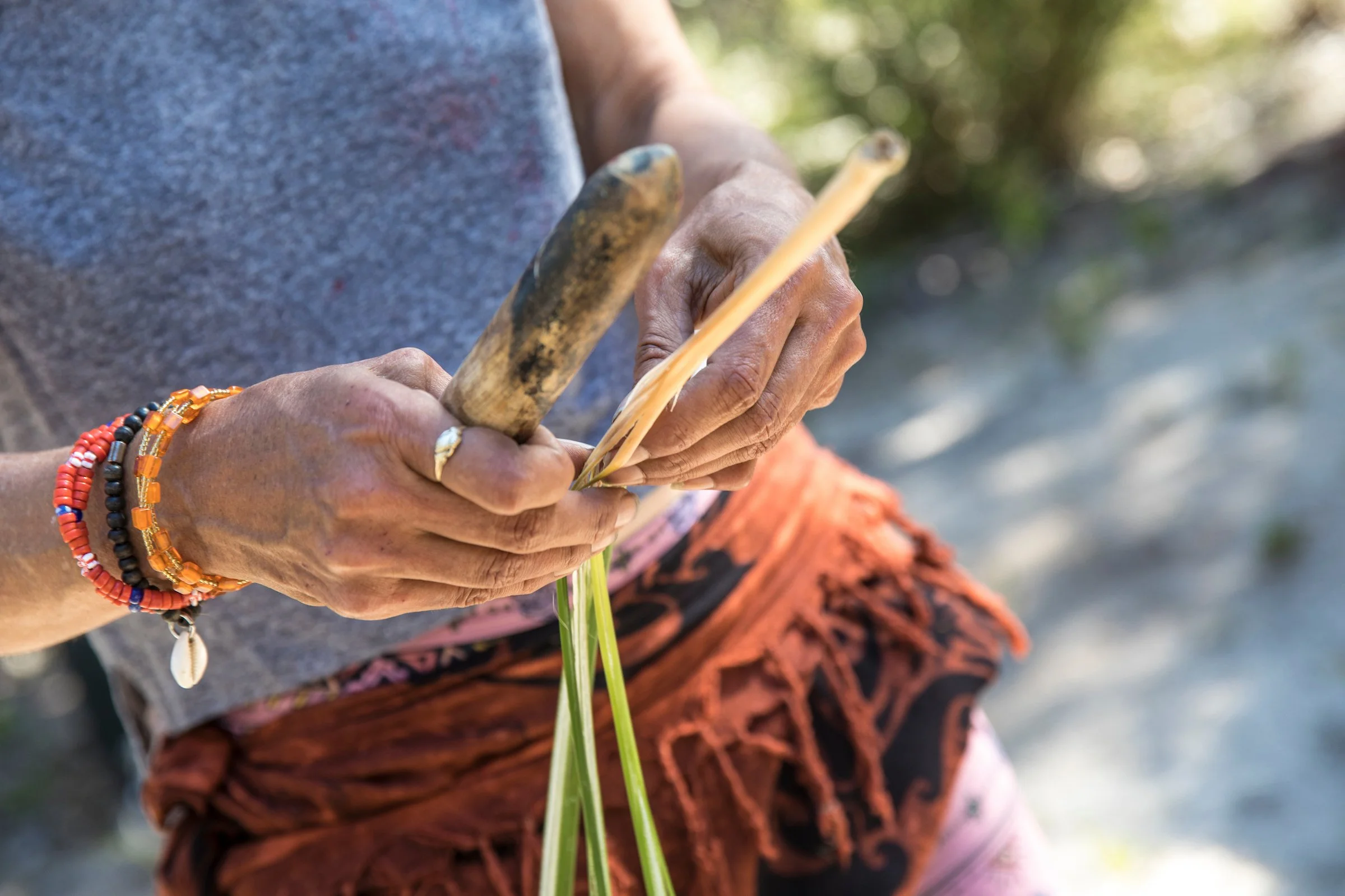 A person holding a tool and working with green plant or grass outdoors, wearing colorful bracelets and a patterned skirt or pants.