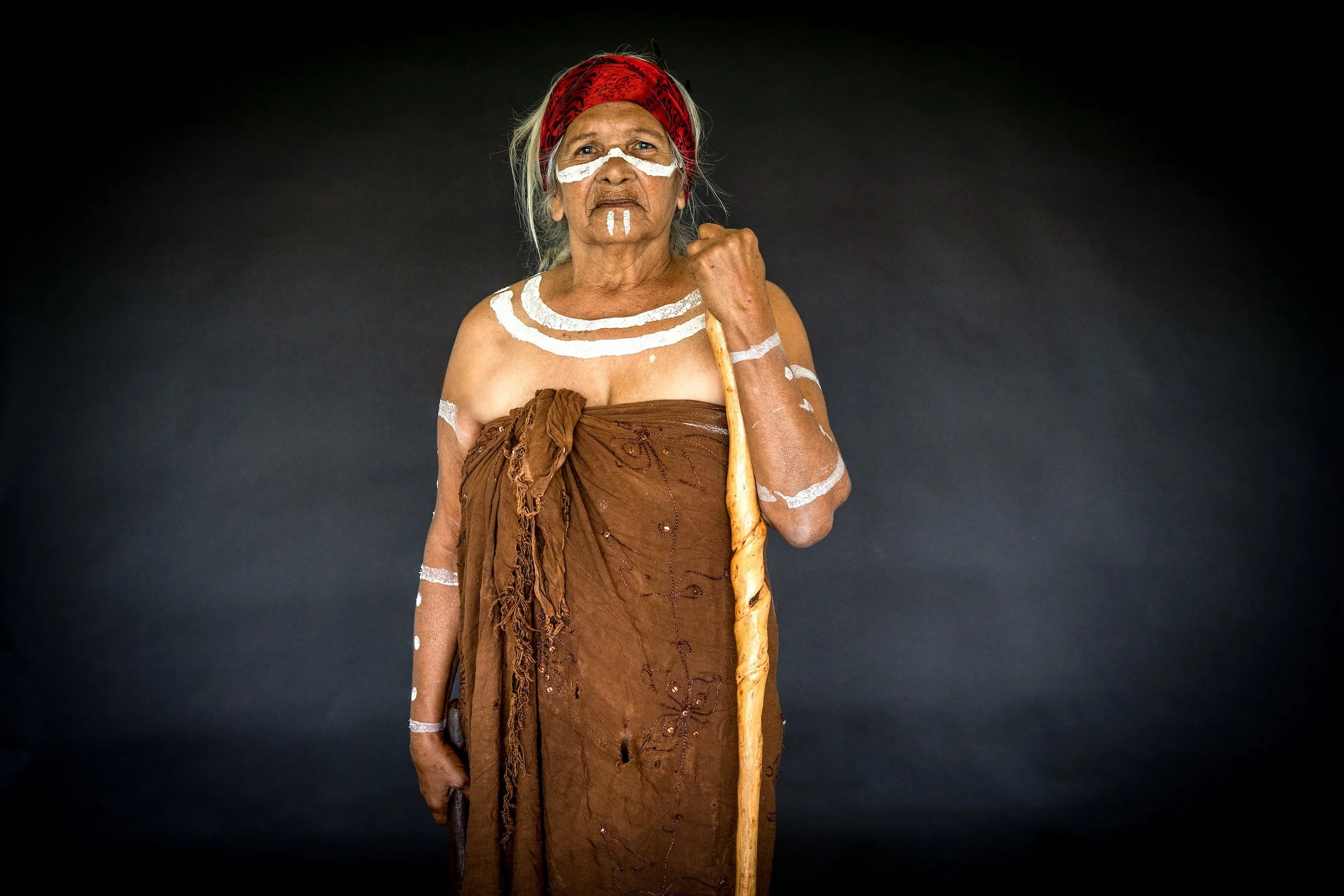An elderly indigenous woman dressed in traditional attire, holding a wooden staff and wearing red and white face paint, standing against a dark background.