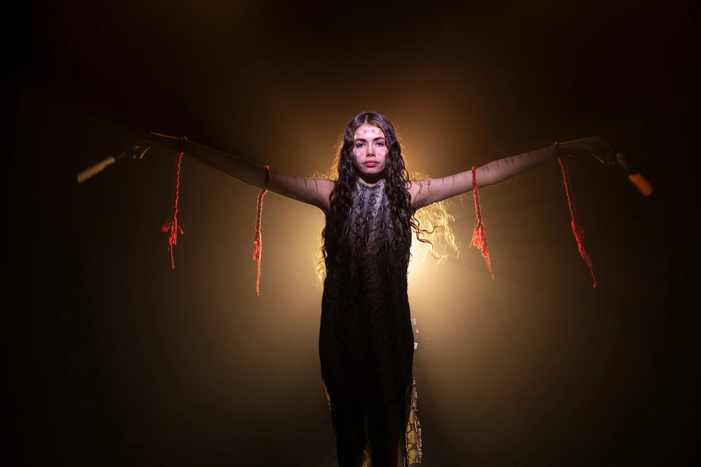 A woman with long curly hair and face paint stands with arms outstretched, holding a long stick with red yarns hanging from it, backlit by a yellowish light in a dark setting.