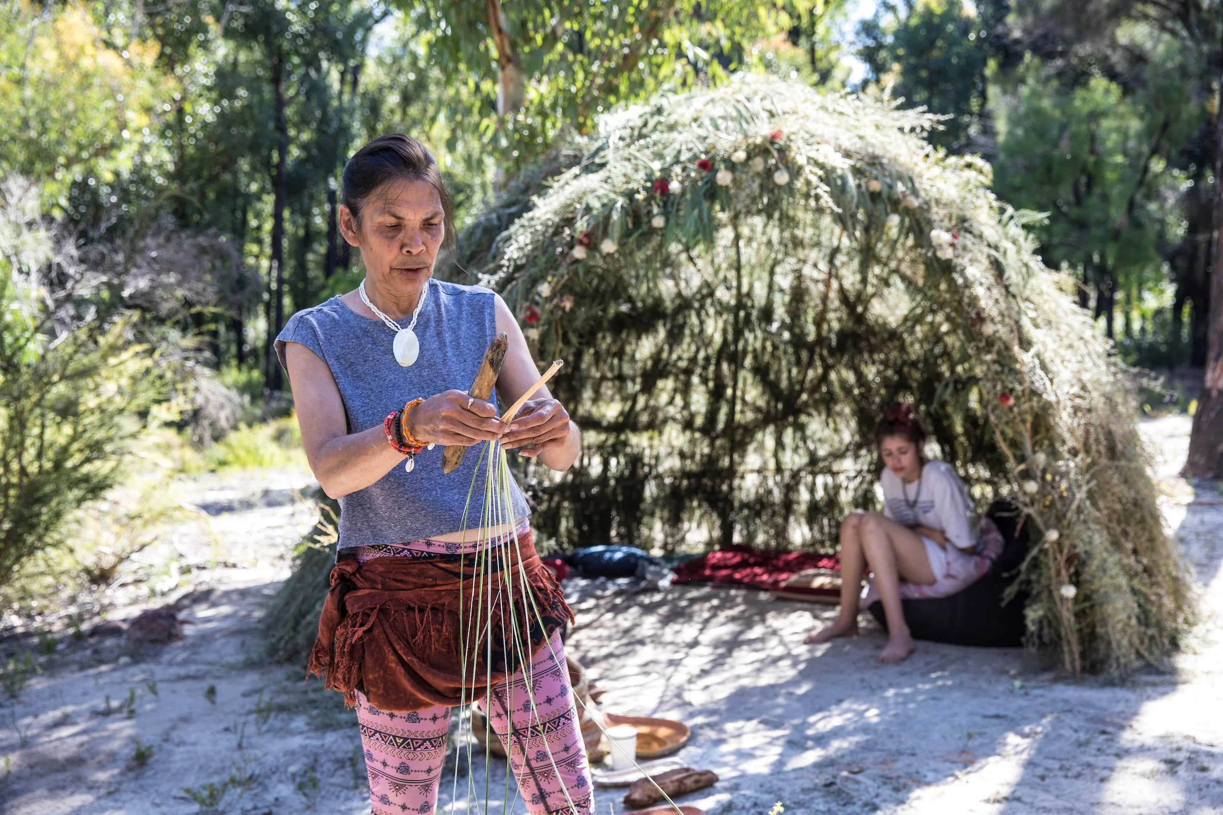 A woman with a necklace and colorful clothing weaving with natural fibers outdoors under a tree, with a young girl sitting nearby.