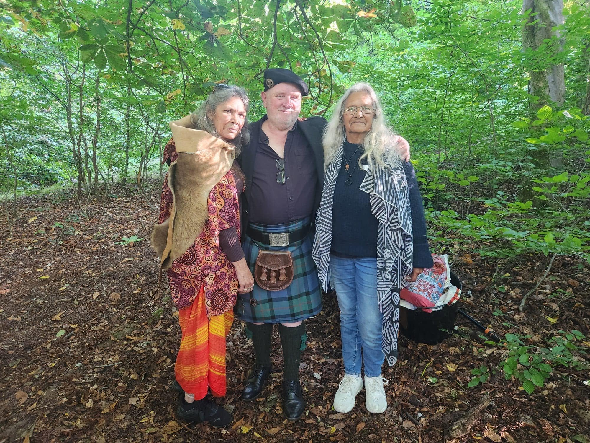 Three people standing together in a lush green forest, posing for a photo. An older woman with long gray hair and glasses is on the right, a man with a beard and wearing a bush hat in the middle, and an older woman on the left with gray hair and a co