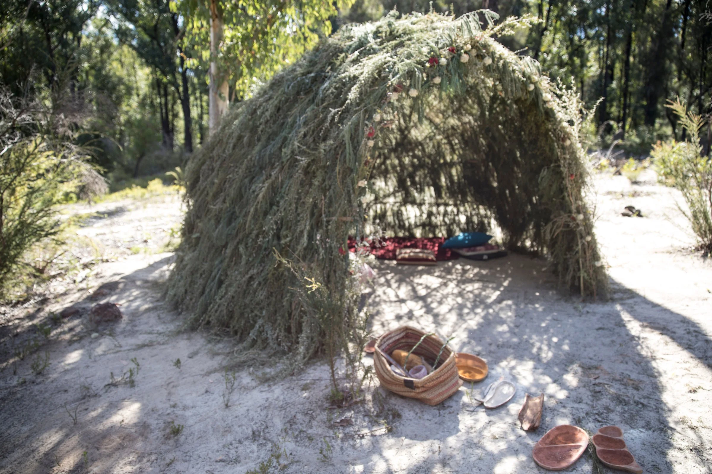 A natural shelter made of branches and foliage in a wooded outdoor setting, with various items including a woven bag, sandals, and blankets placed around the entrance.