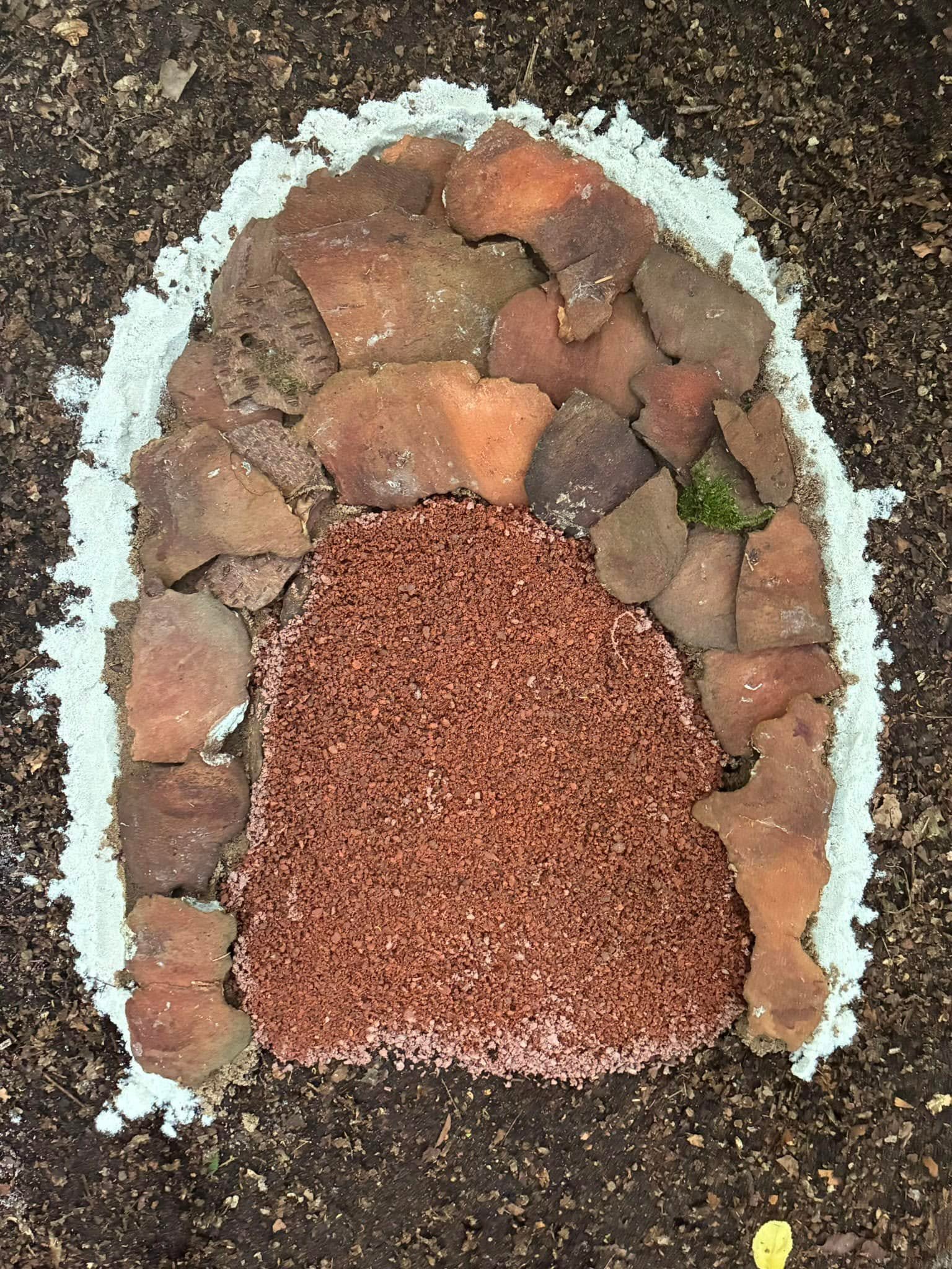 A fire pit made of bricks, with some rocks and a small patch of moss inside, surrounded by dirt and gravel.