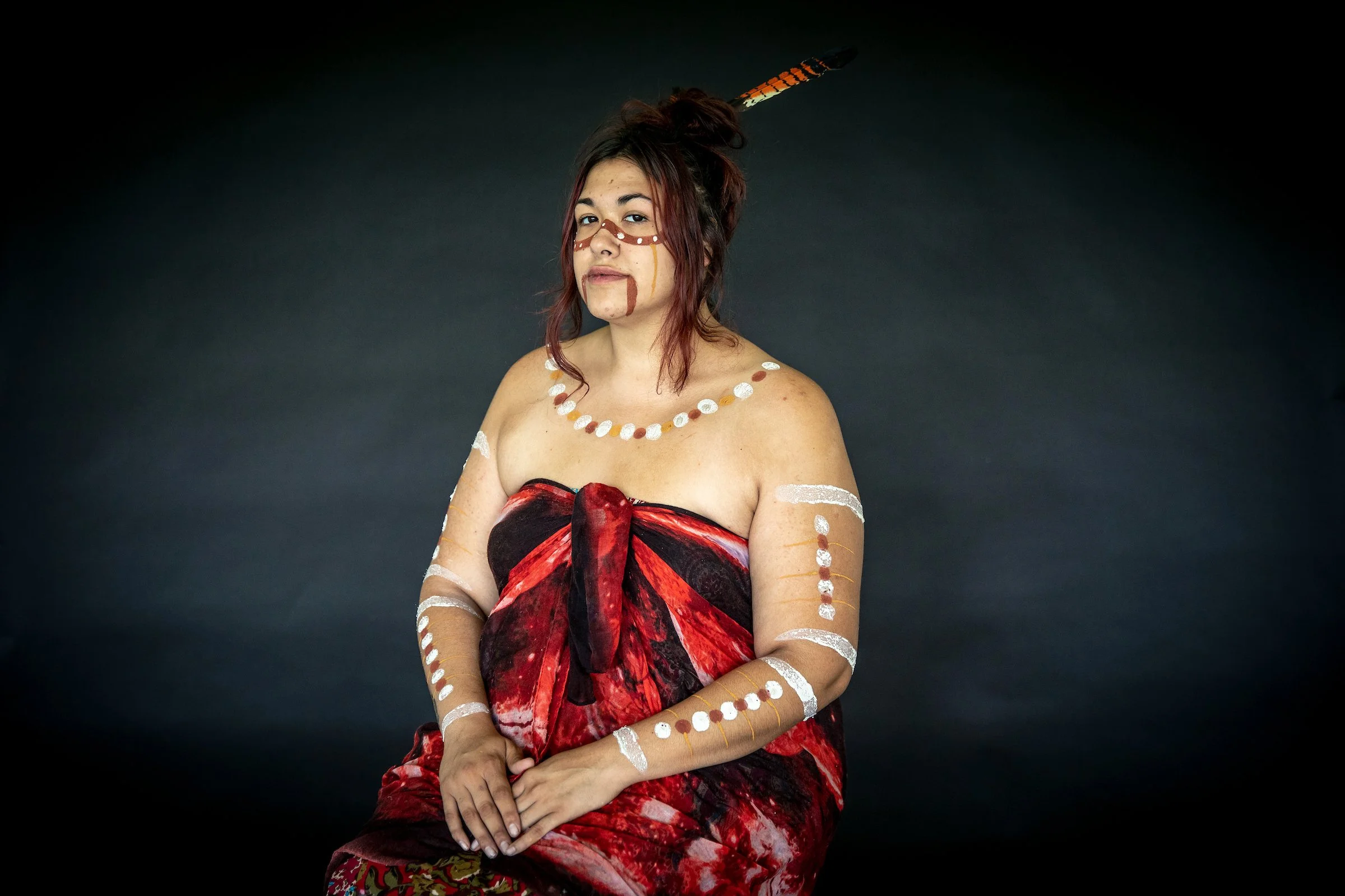 Woman dressed in traditional indigenous attire with body paint, sitting against a black background.