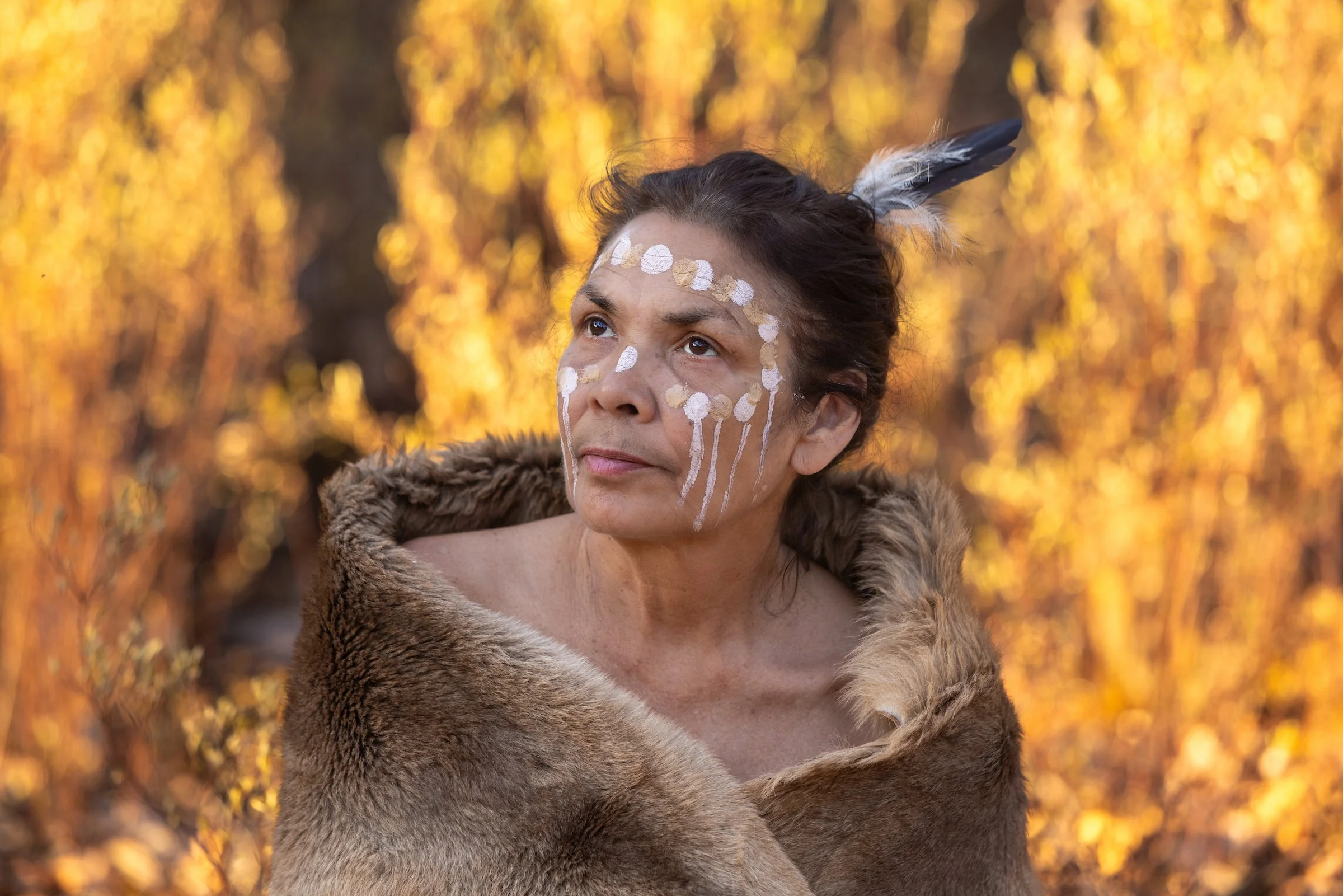 A woman with face paint and feathers in her hair, wearing fur, looking upward in front of a blurred background of autumn trees.