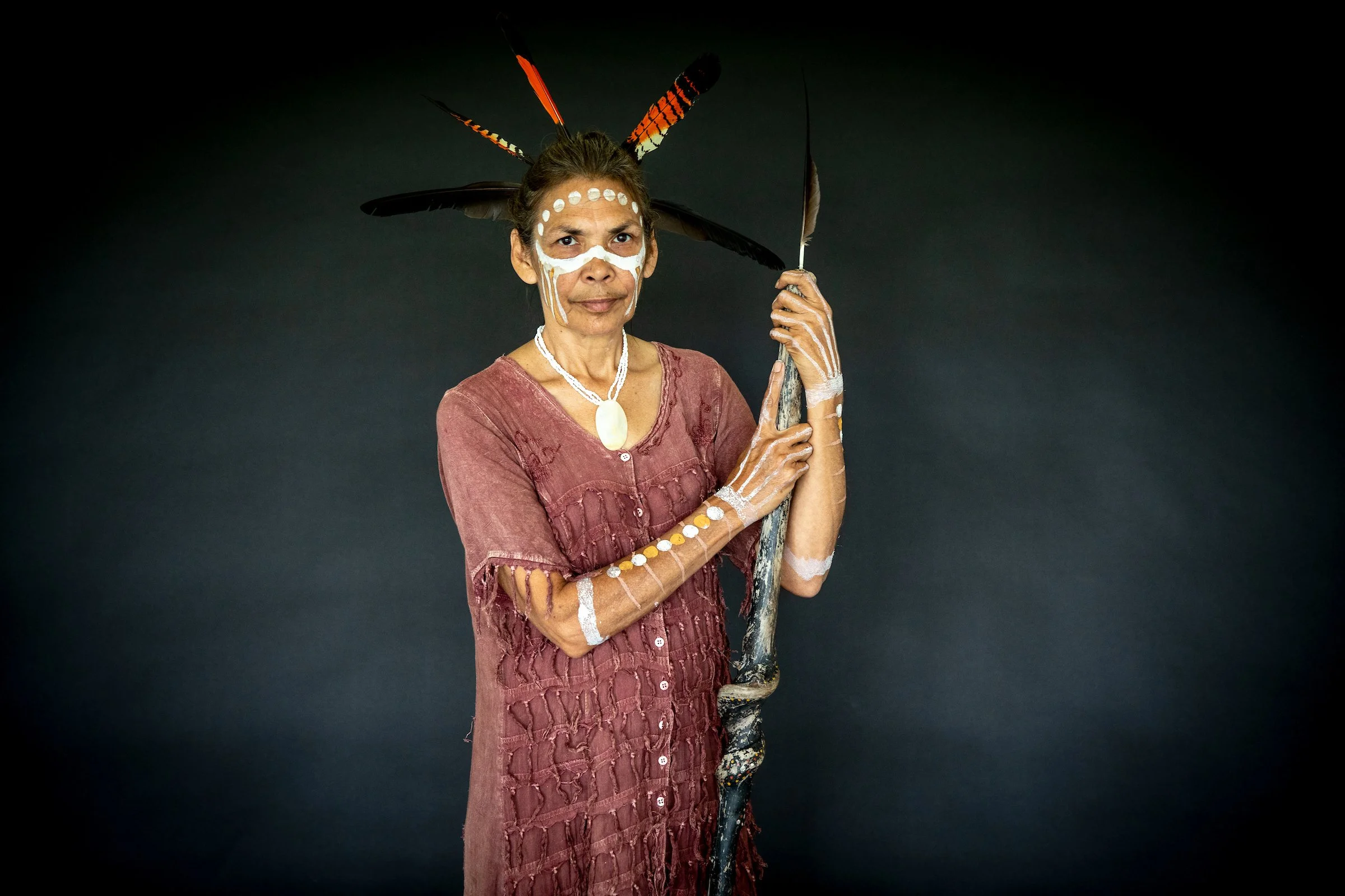 A woman dressed in traditional indigenous attire with face and body paint, wearing a headpiece made of feathers, holding a staff, and standing against a dark background.