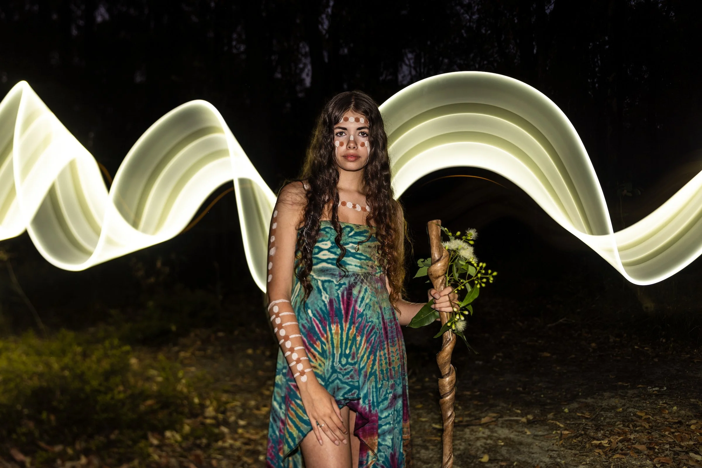 A young woman with long curly hair and face paint standing outdoors at night, holding a decorated staff and flowers, with light trails behind her.