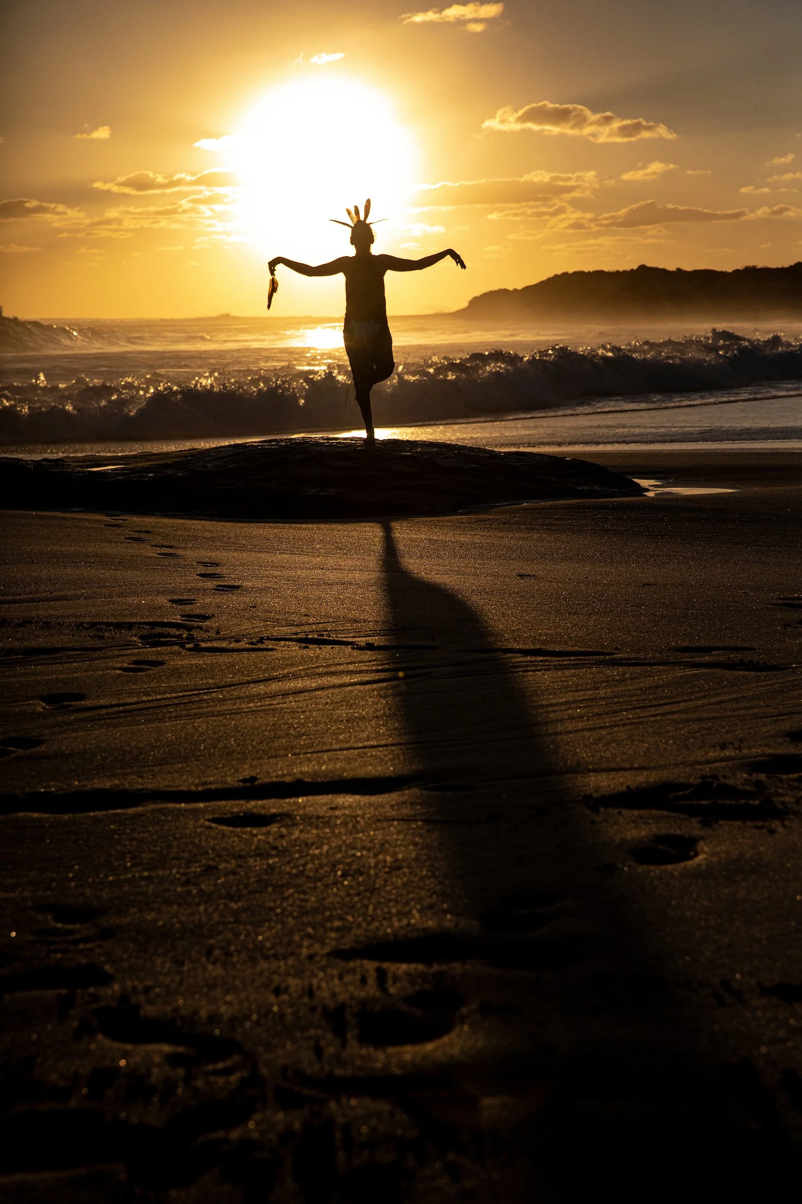 Person standing on a beach at sunset, wearing a headband with feathers, arms outstretched, facing the sun.