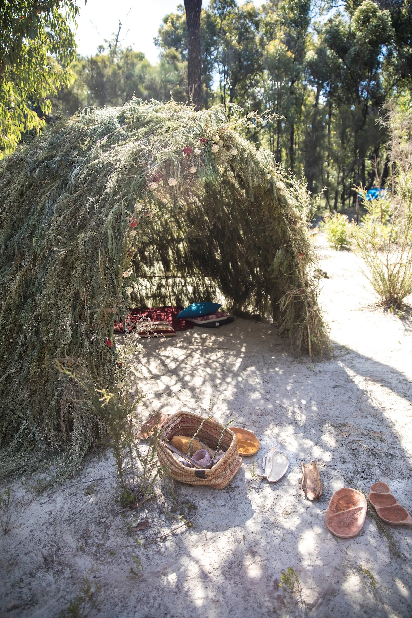 A small outdoor altar or shrine on a sandy ground, made with a natural arch structure covered in greenery and flowers. Inside the altar, there are cushions and offerings, including pottery bowls, baskets, and other small objects. It is set in a woode