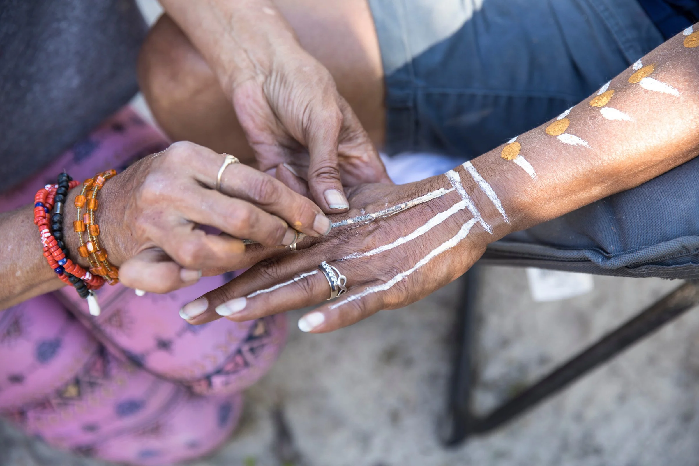 A person with darker skin applying white paint or chalk lines on another person's arm, who is wearing multiple colorful beaded bracelets, a floral shirt, and the background shows a person wearing a gray shirt and denim shorts.