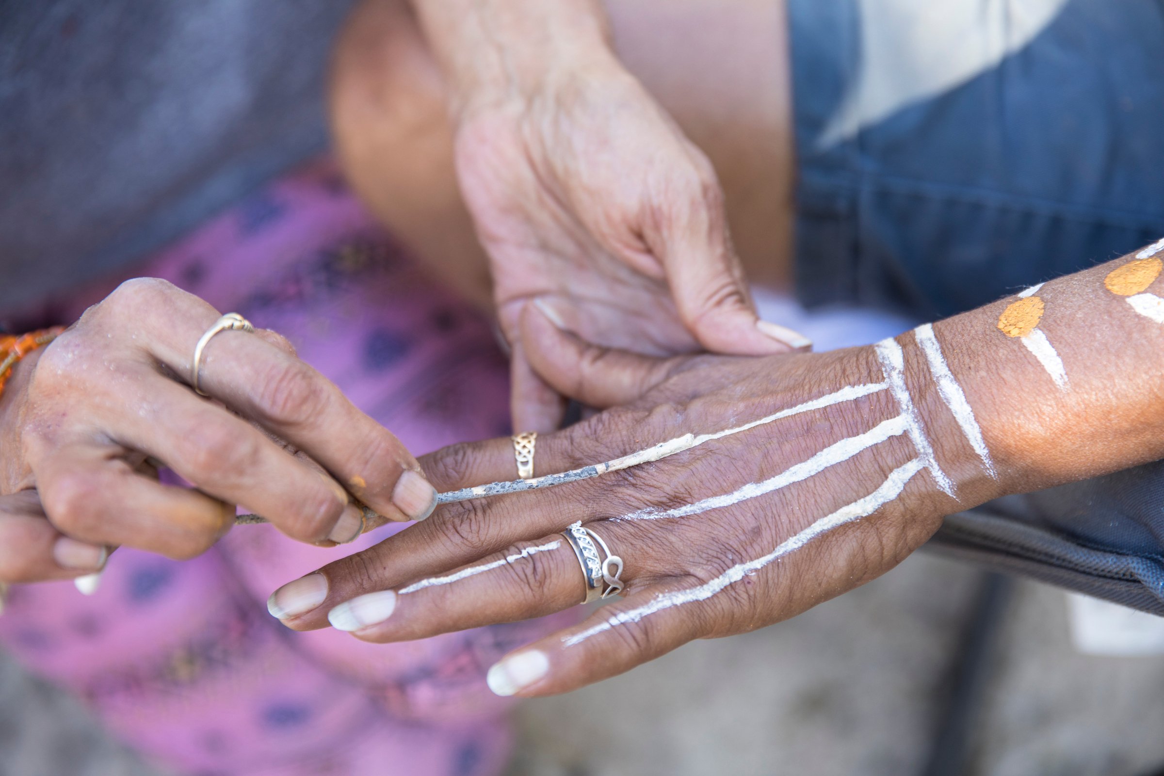 Close-up of elderly hands applying white and yellow traditional paint or chalk design on a person's arm and hand, with people wearing rings and colorful clothing in the background.