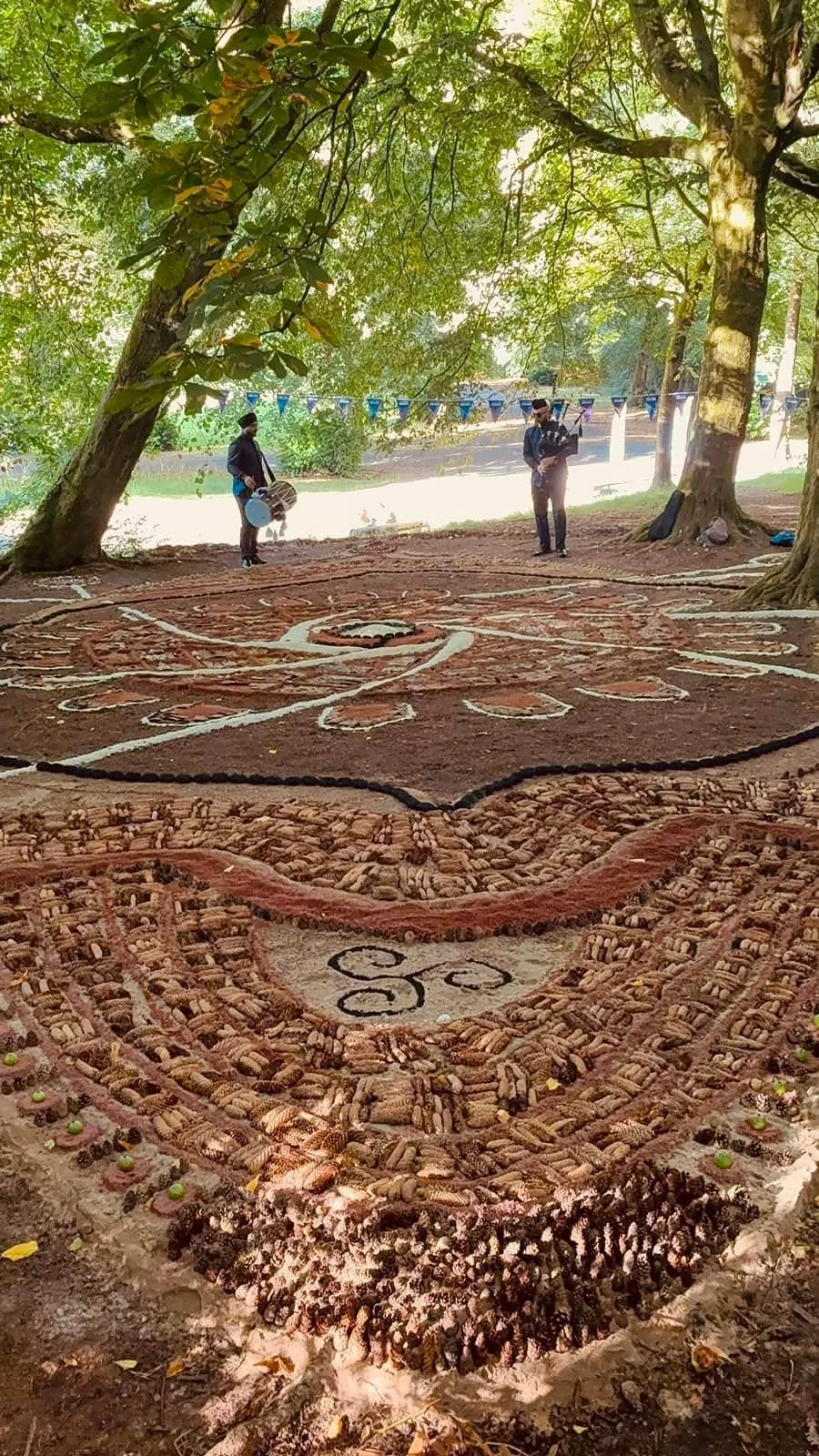 Colorful sand mandala art on ground in a park with trees and two musicians nearby.