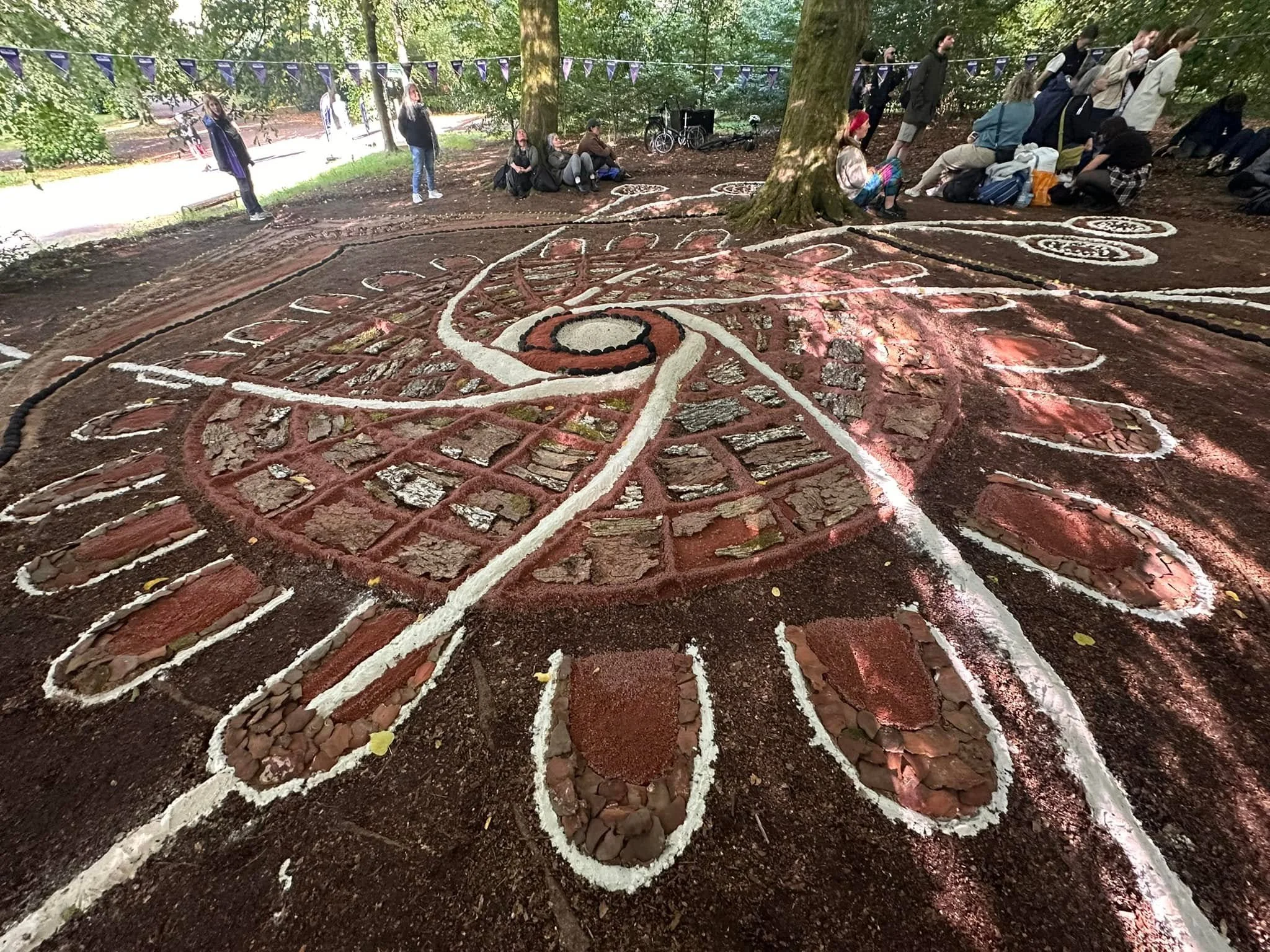 People sitting and standing around a spiral chalk drawing on the ground in a wooded outdoor area.