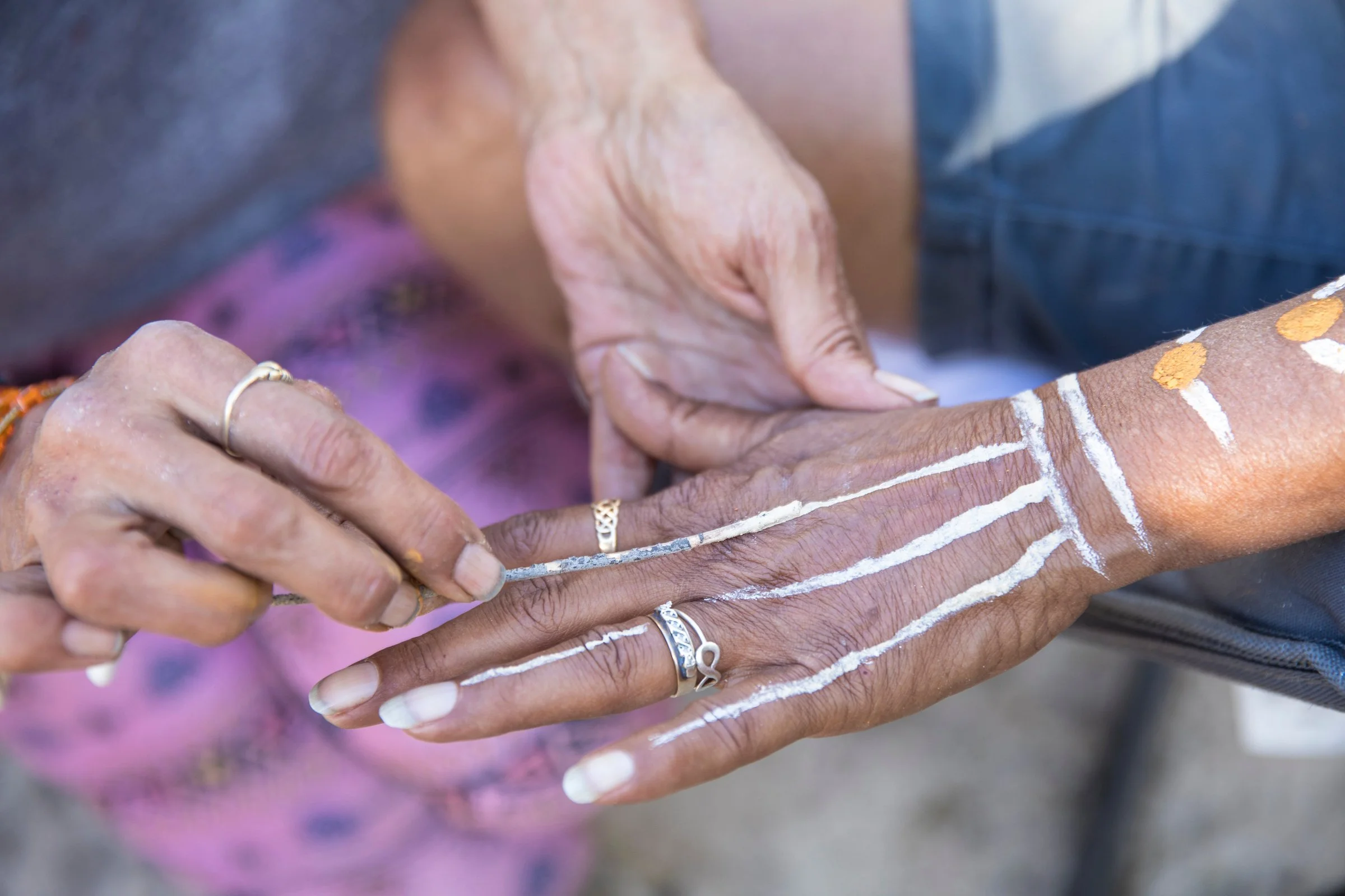 Close-up of two elderly hands with white painted lines and traditional jewelry, one hand assisting the other. The background includes a person wearing a gray shirt and blue jeans.