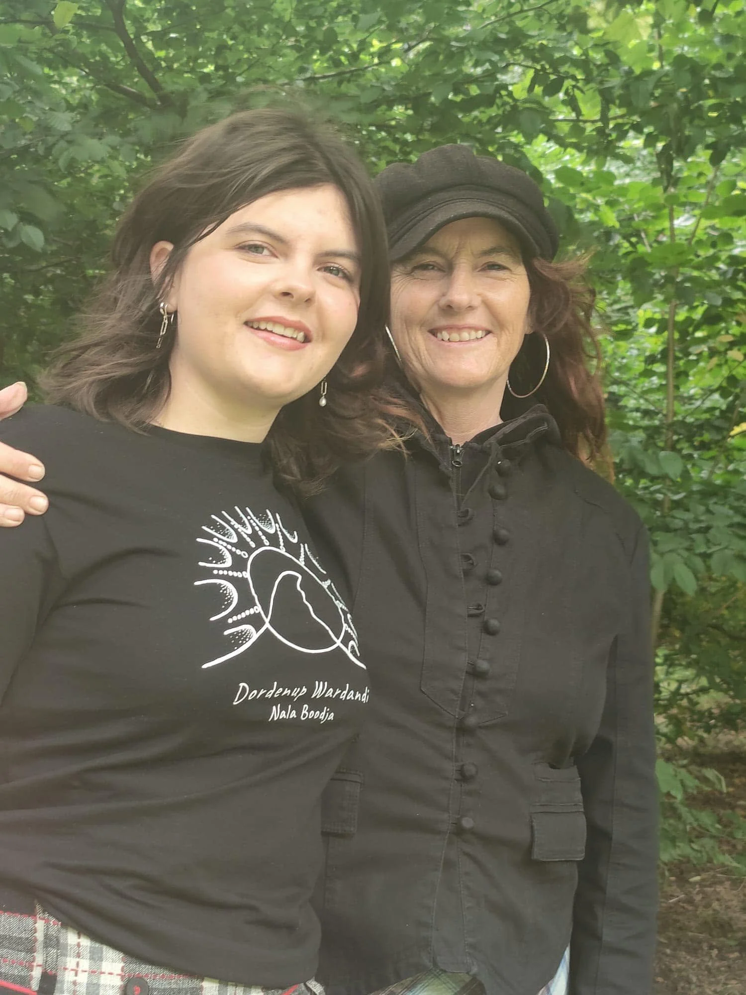 Two women smiling and standing close together outdoors among green trees. The woman on the left has dark, shoulder-length hair, earrings, and is wearing a black shirt with white text. The woman on the right has curly brown hair, an African American-s
