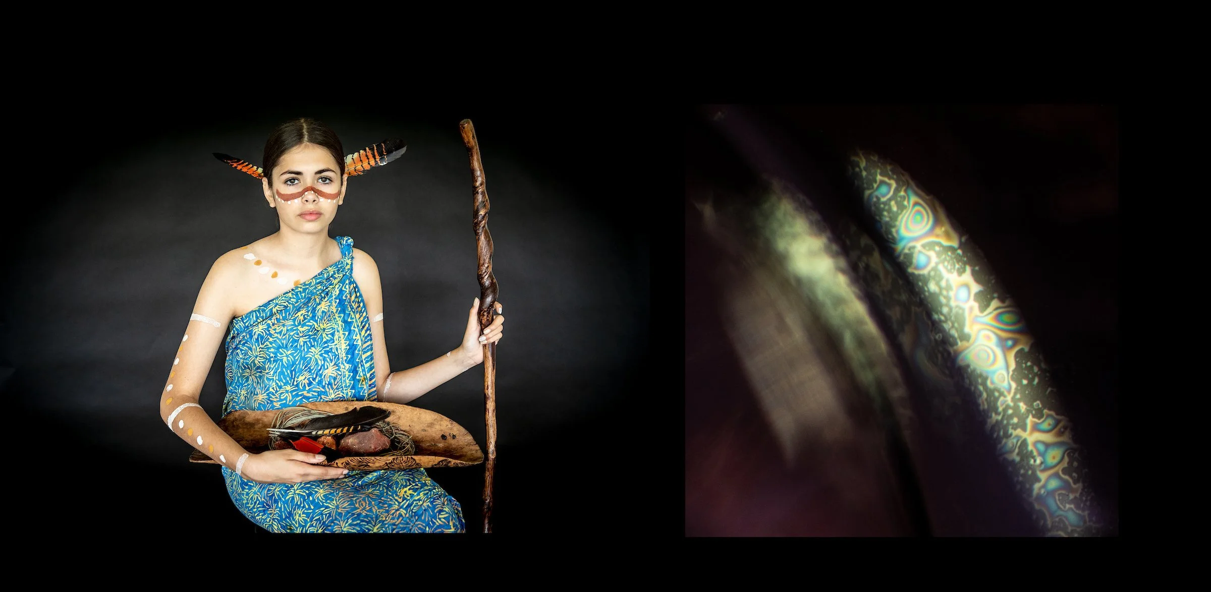 A young woman dressed in traditional indigenous clothing and face paint, holding a staff and a bowl with natural objects. She has butterfly and feather accessories and painted dots and lines on her arms and face. The background is dark.