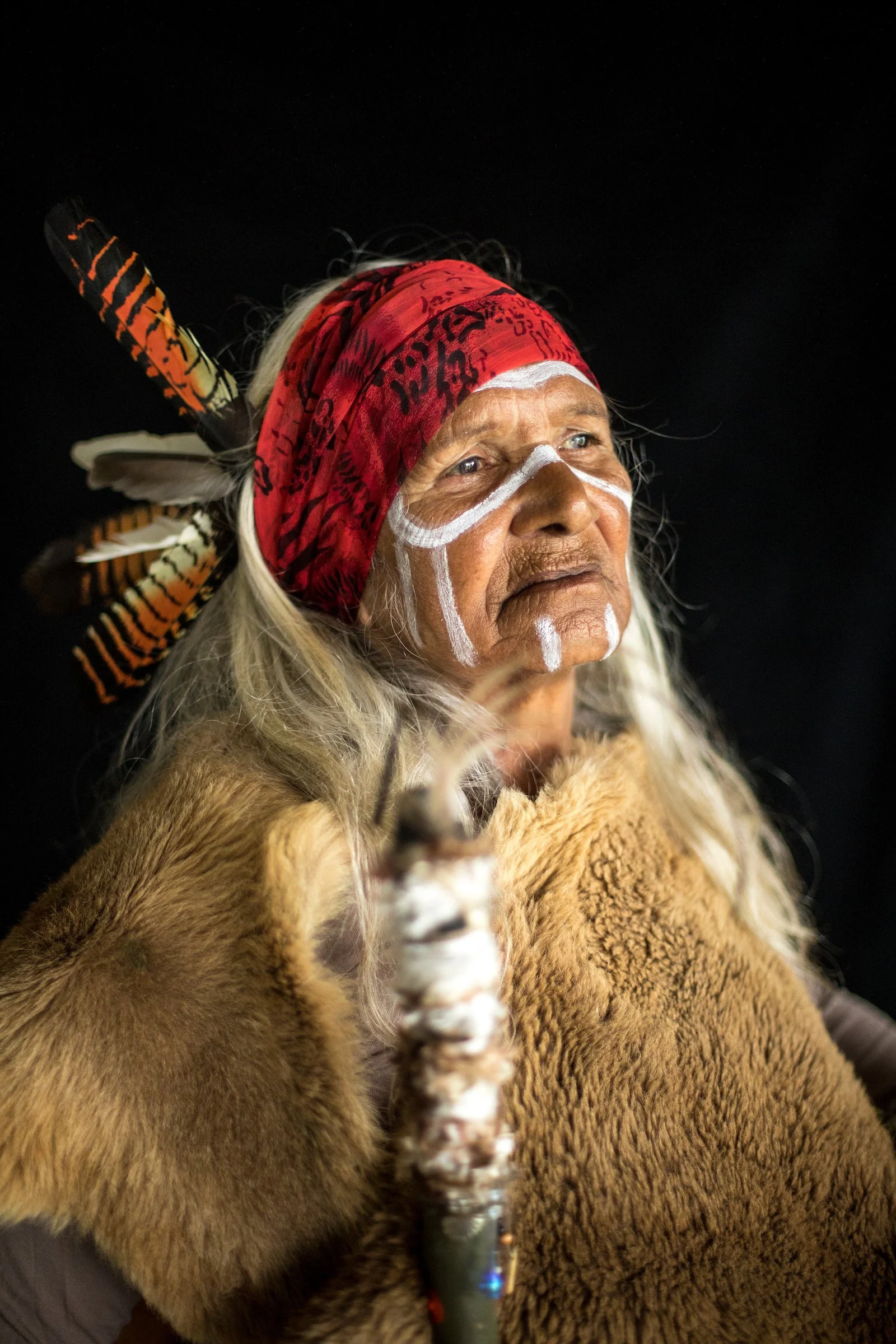 An Indigenous man with face paint, wearing a red bandana with black writing and feathers, holding the reins of a brown animal, likely a dog or a domestic animal, against a black background.