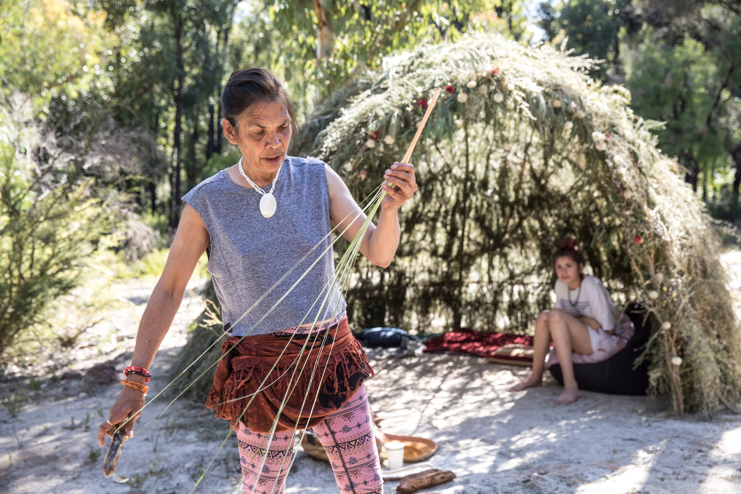 A woman with dark hair tied back, wearing a gray sleeveless top, patterned pink pants, and jewelry, is holding a stick or rope outdoors in a natural setting with trees and greenery. Another person is sitting on the ground in the background.