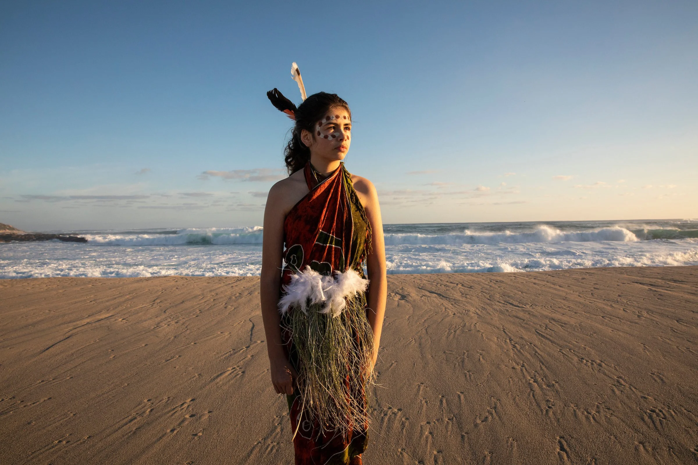 A woman dressed in traditional Native American attire standing on a sandy beach with ocean waves and a clear sky in the background.