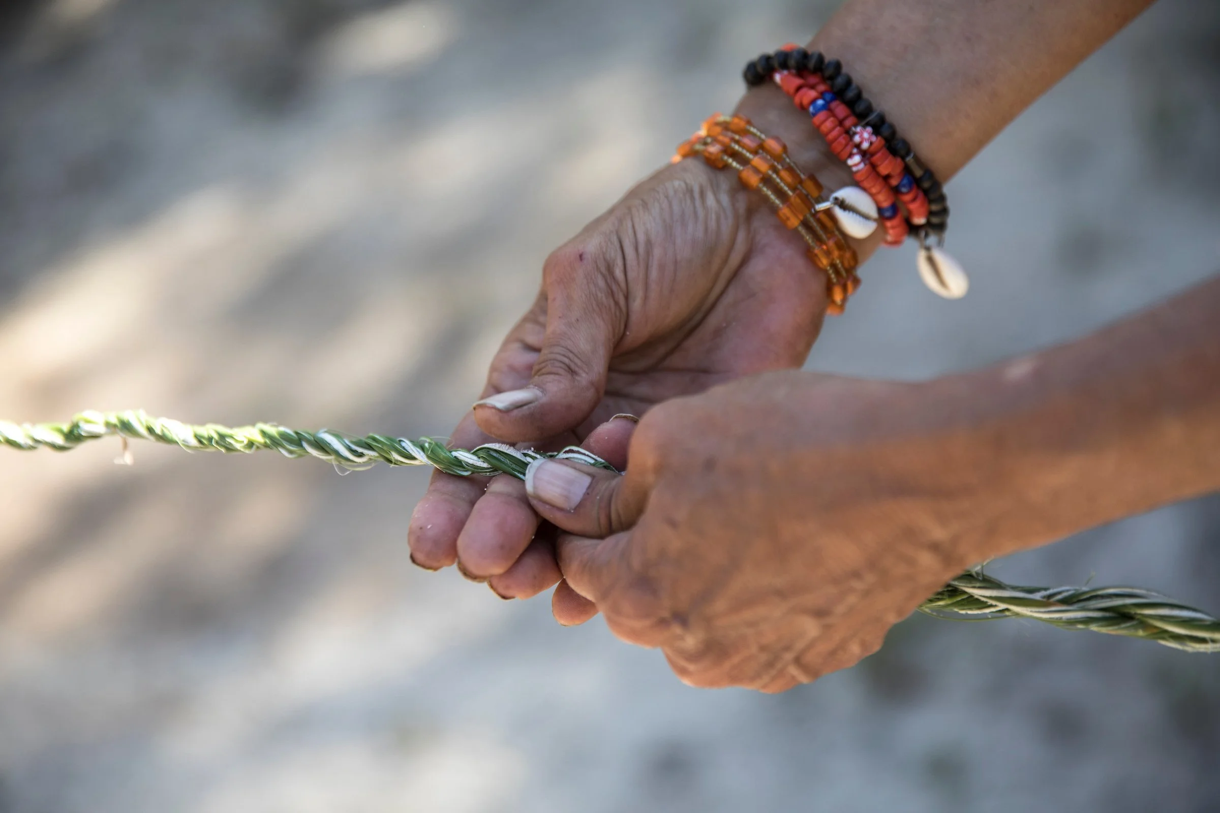 Close-up of two hands holding a woven craft item, with one hand wearing colorful beaded bracelets and shell charms, set against a blurred outdoor background.