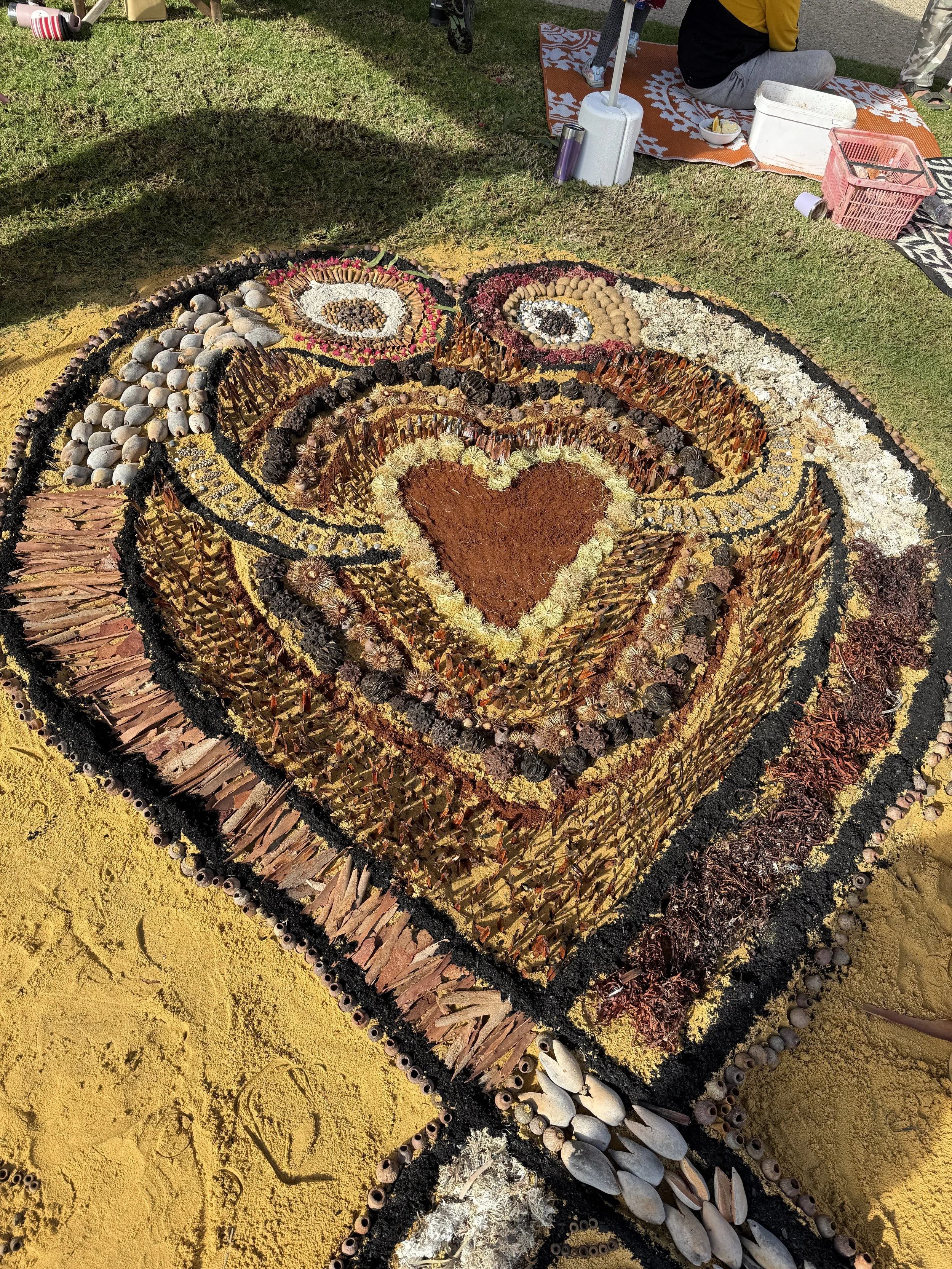 Sand art design on the ground featuring concentric patterns and a heart shape at the center, decorated with seashells, stones, and natural materials.