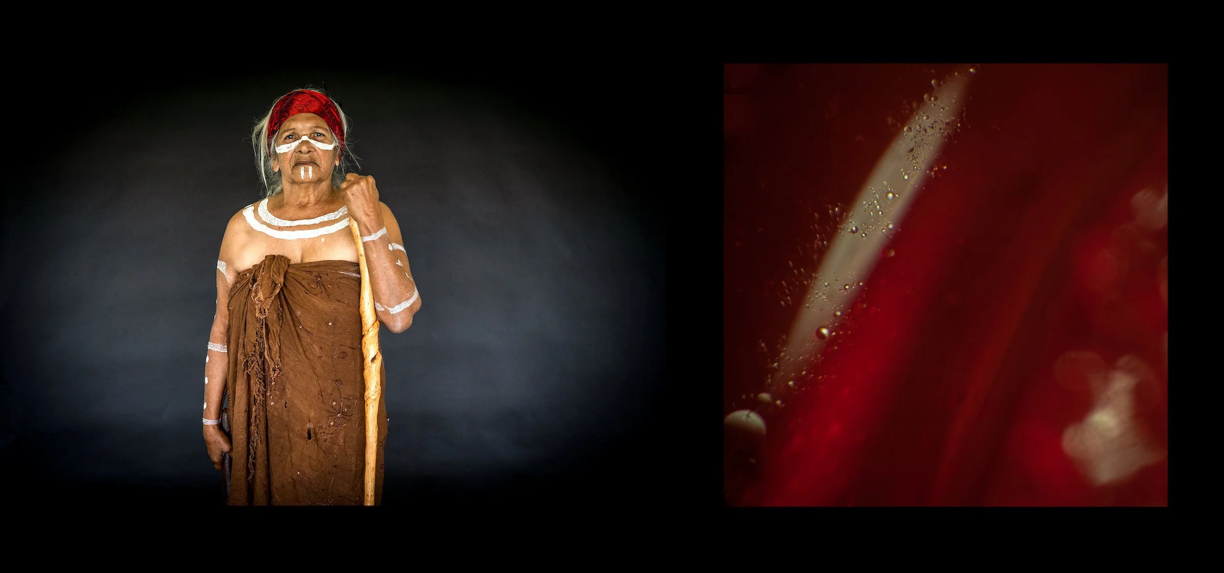 An elderly Indigenous woman dressed in traditional attire, with white face paint and a red headscarf, holding a wooden staff, standing against a plain black background.