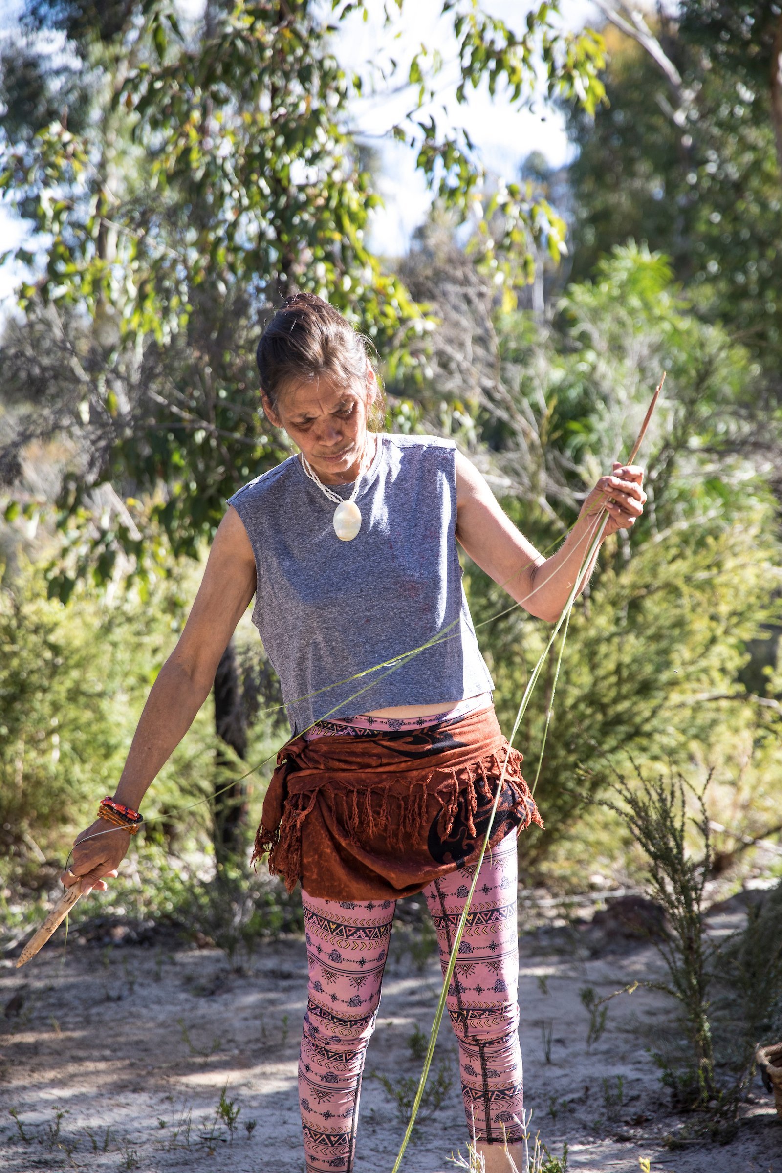 A woman standing outdoors among trees, rope in hand, wearing a sleeveless top, patterned pants, and a large shell necklace.