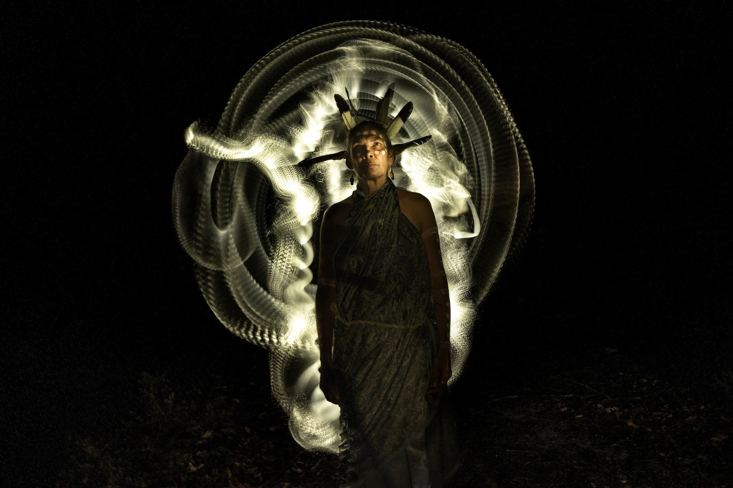 A person dressed in traditional Native American attire, including face paint and feathers, stands in front of a light installation creating intricate light patterns against a dark background.