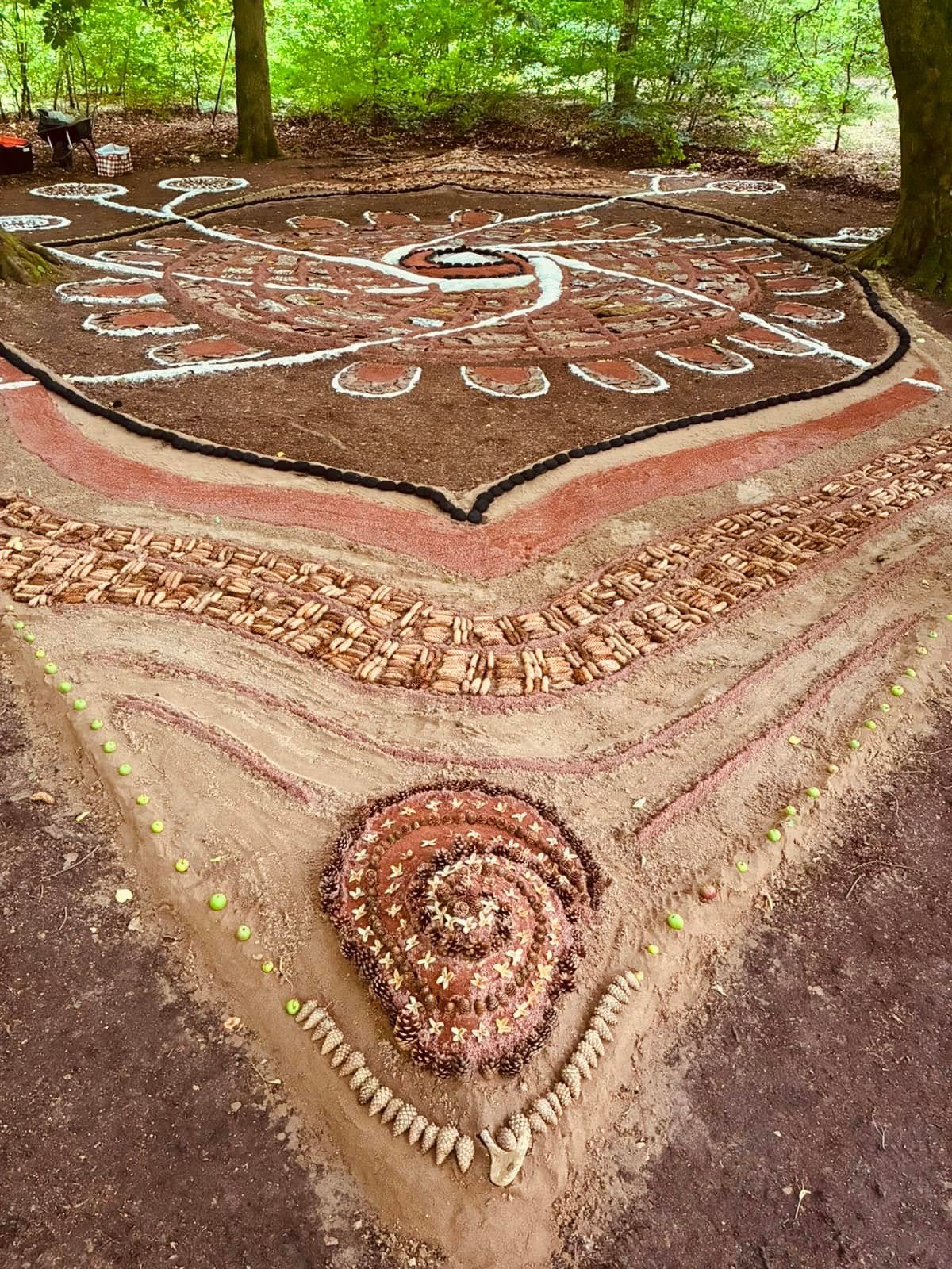 A large outdoor sand art design with spiral and geometric patterns created with shells, stones, and other natural materials, bordered by small green balls, in a wooded area.