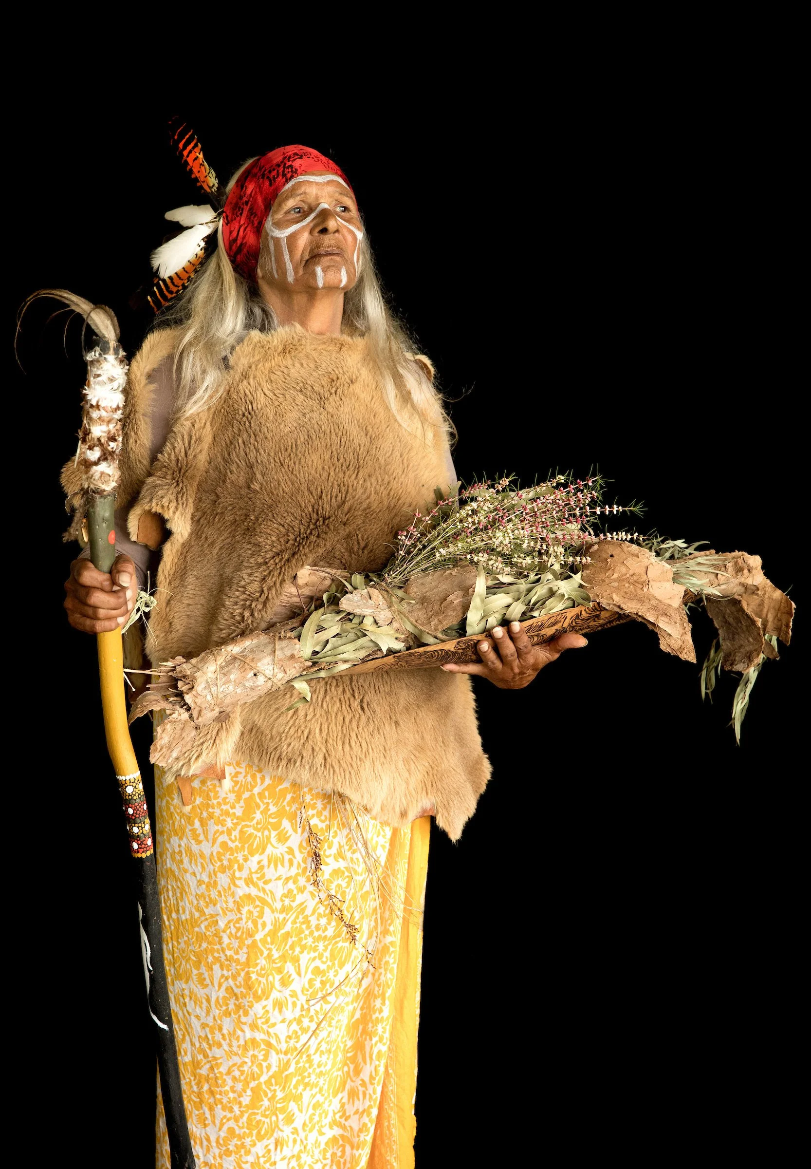 A person dressed in traditional indigenous attire holding a ceremonial item with flowers and foliage, against a black background.